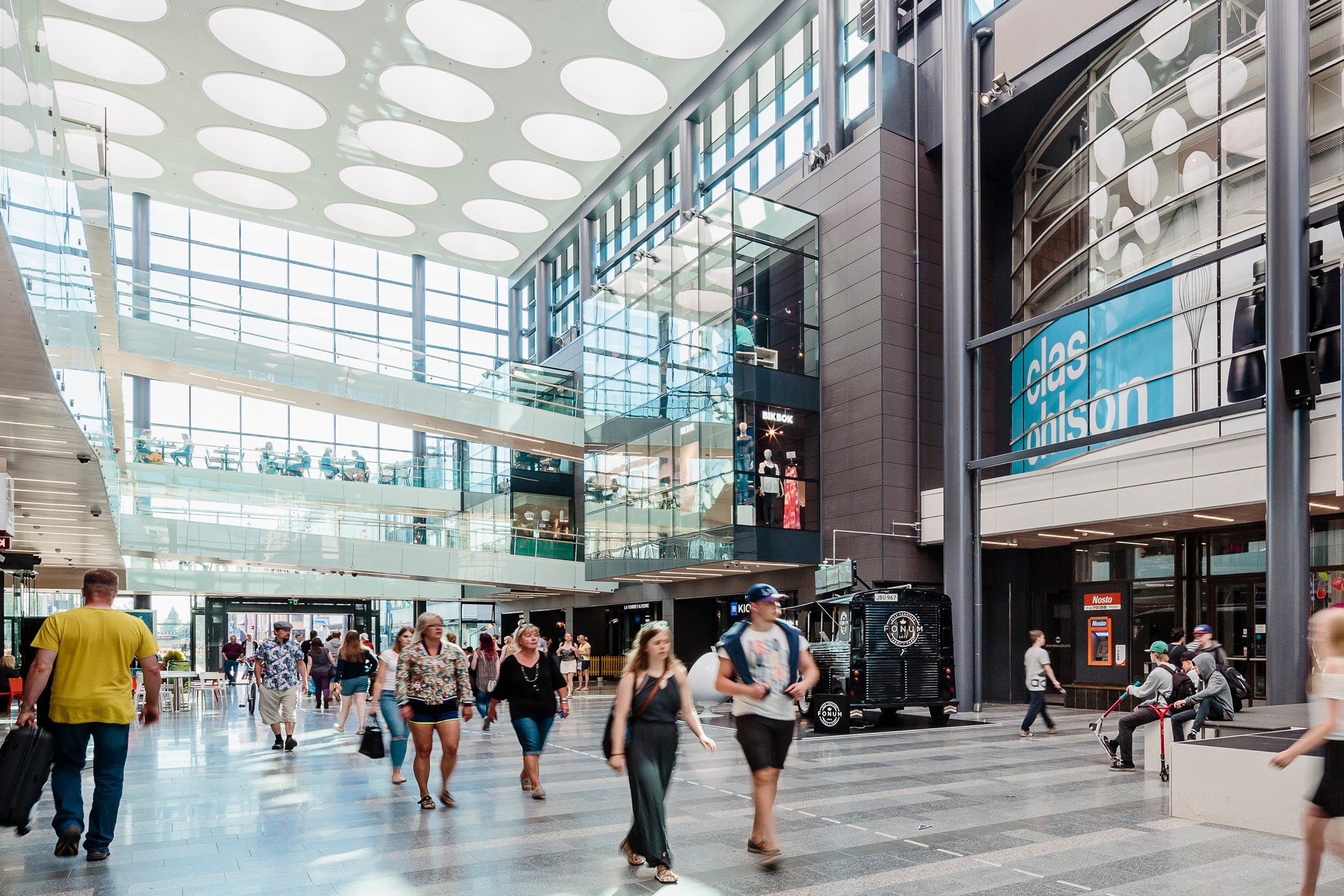 Spacious shopping mall interior with modern architecture, large windows, and people walking by.