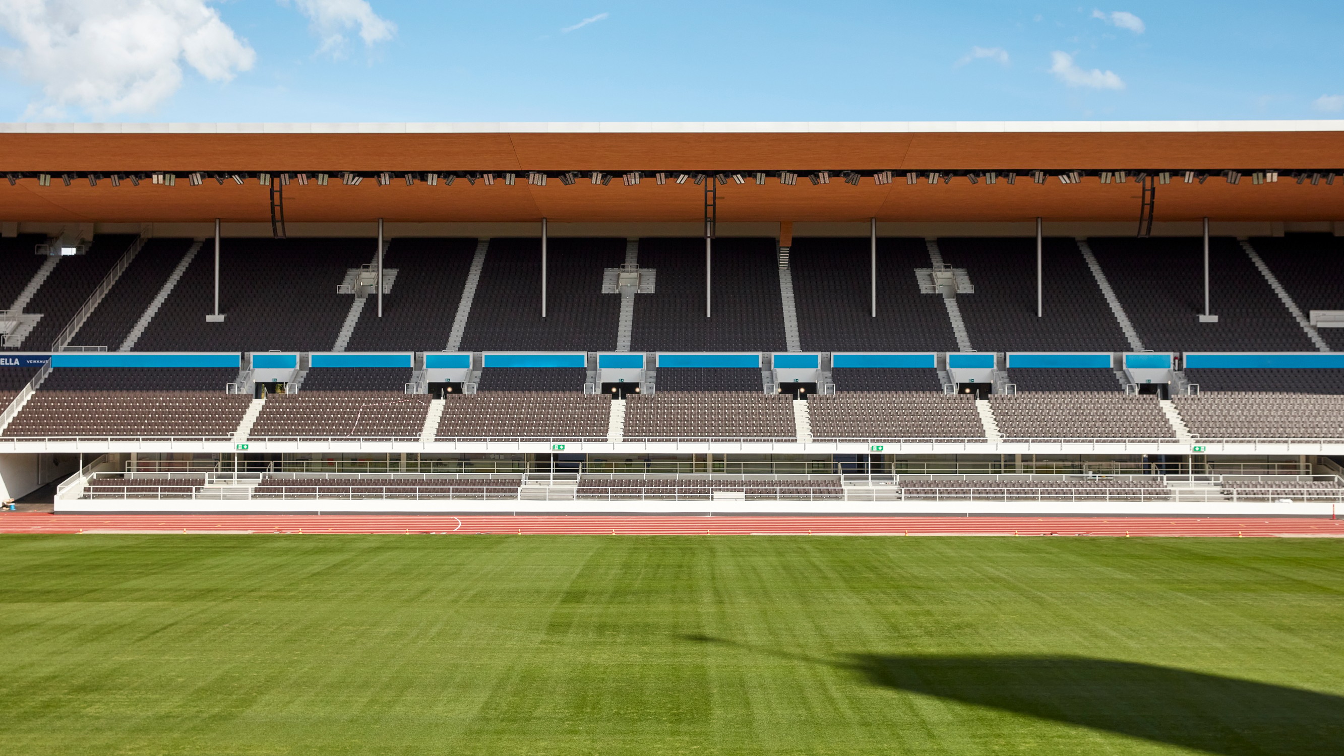 Stadium with wooden roof, tiered seating, and green field under a clear blue sky.