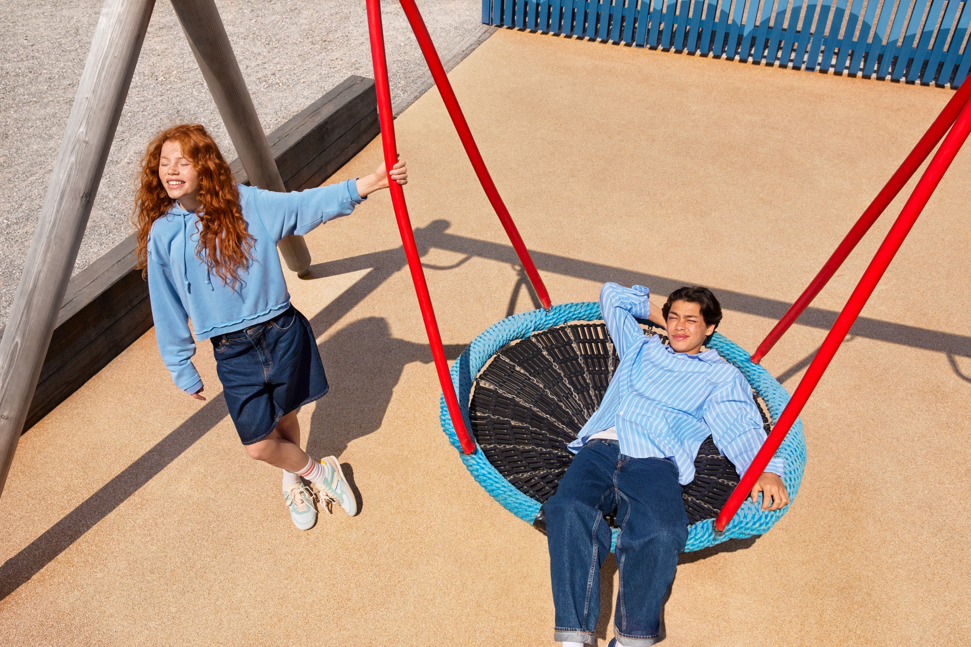 Two children on a playground, one standing and one lounging on a circular swing with red ropes.