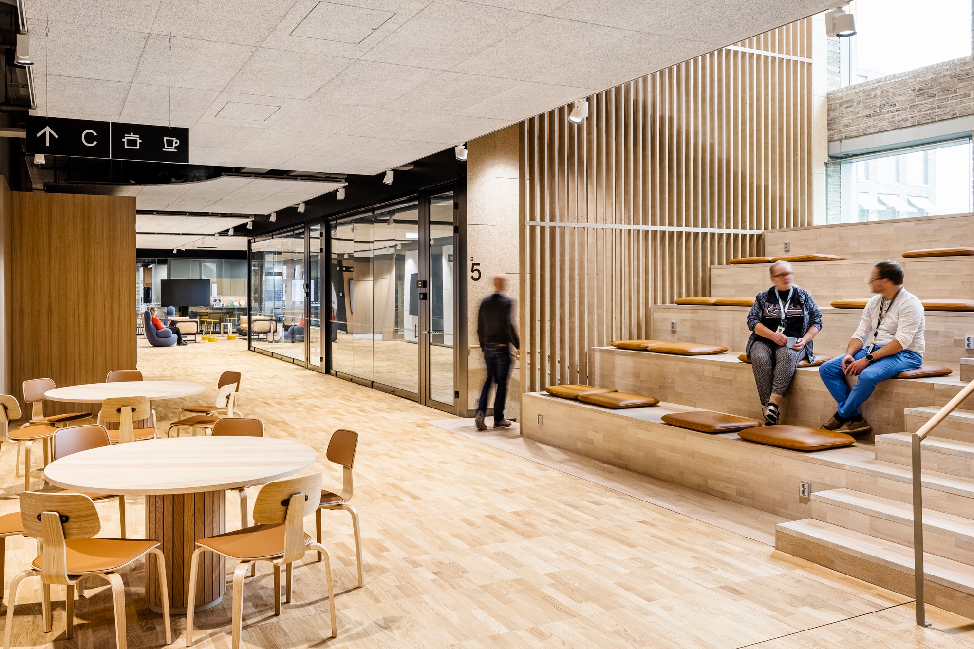 Modern office space with tiered seating, wooden flooring, round tables, and glass partition walls.