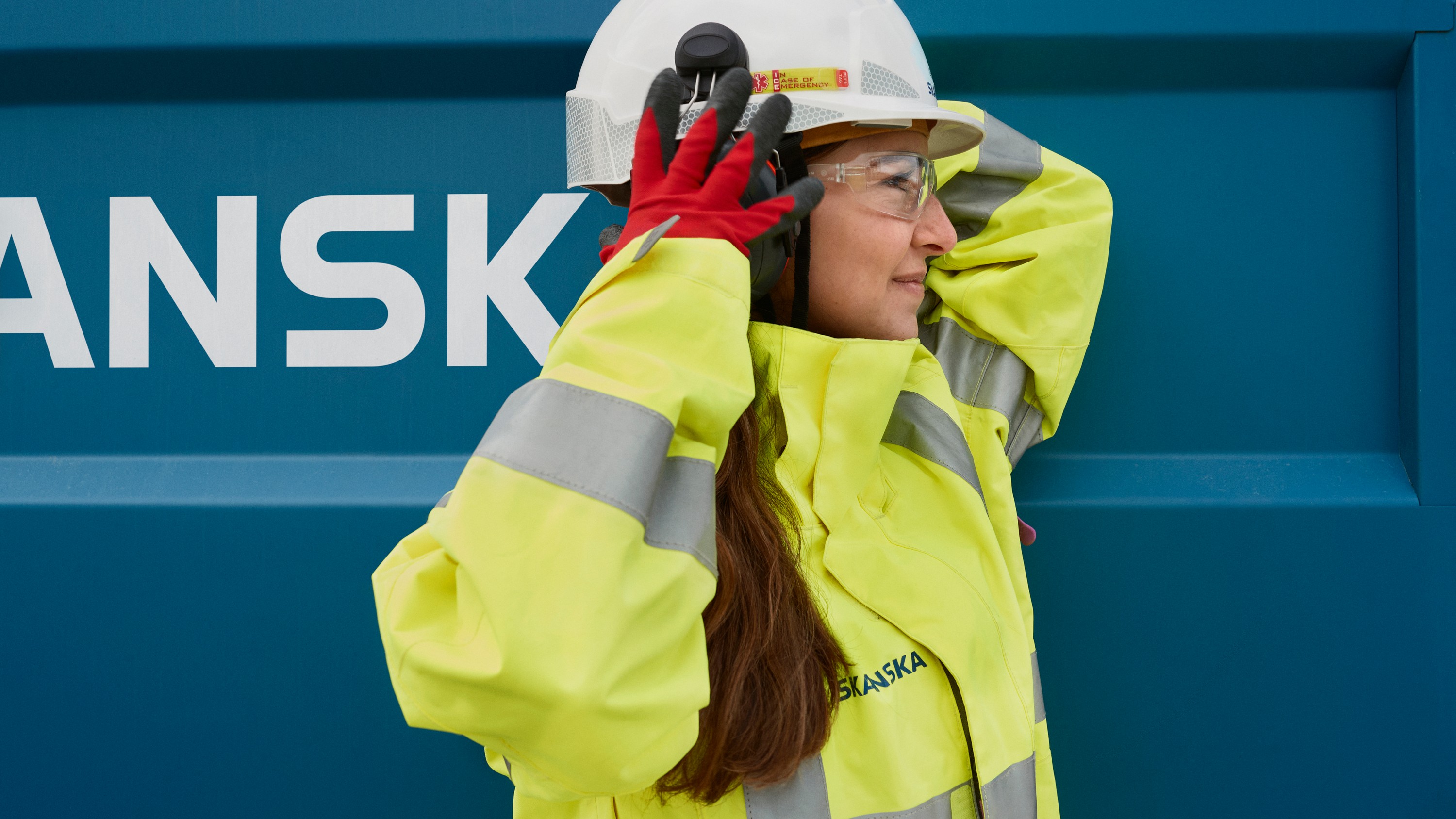 Rakennustyöntekijä näkyvissä olevissa varusteissa säätää kypärää Skanskan kontin edessä.