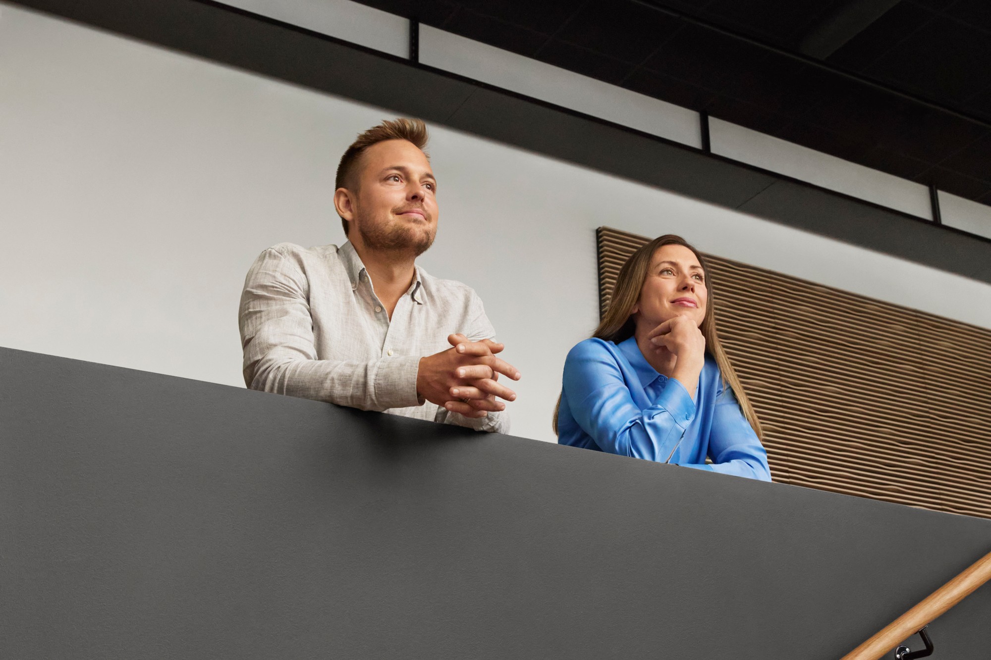 Two thoughtful people looking ahead while leaning on a modern indoor balcony railing.
