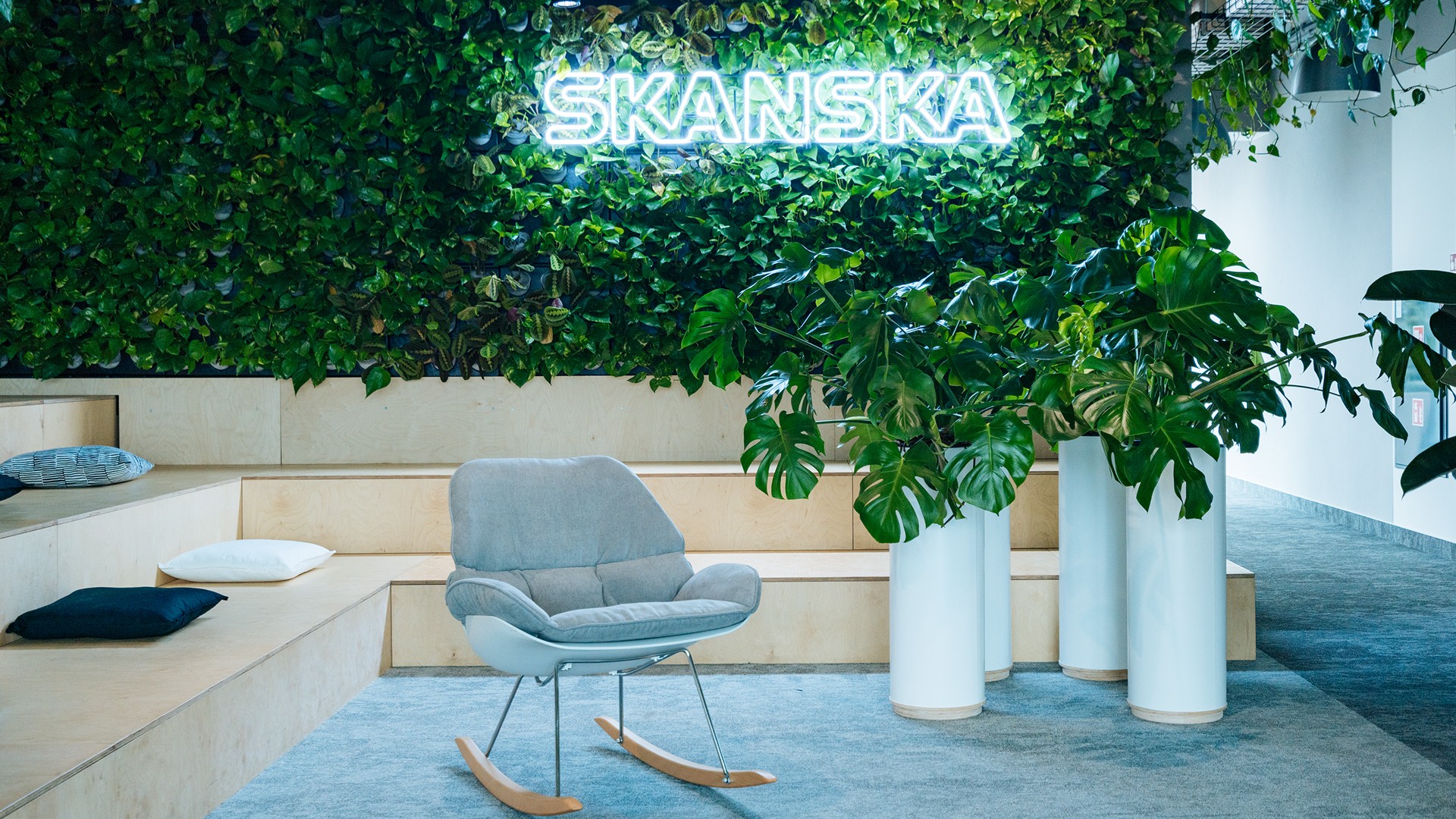 Moderni toimistolounge vihreällä Skanskan pääkonttorissa Moderni toimistolounge, jossa Skanskan logo, vihreä seinä, keinutuoli ja ruukkukasveja tunnelmaa varten.
