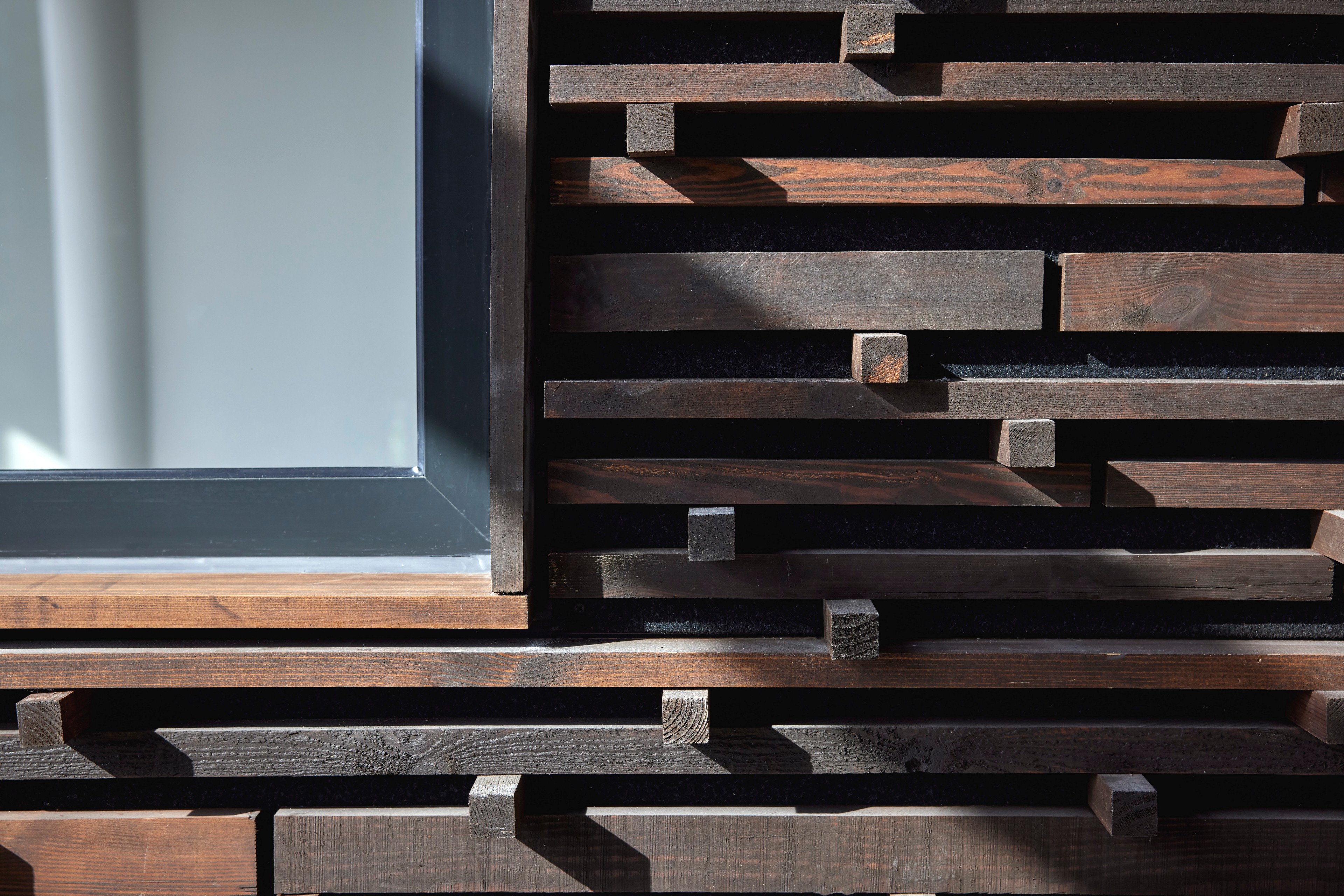 Close-up of a modern wooden slat wall with varied horizontal beams and a window frame.