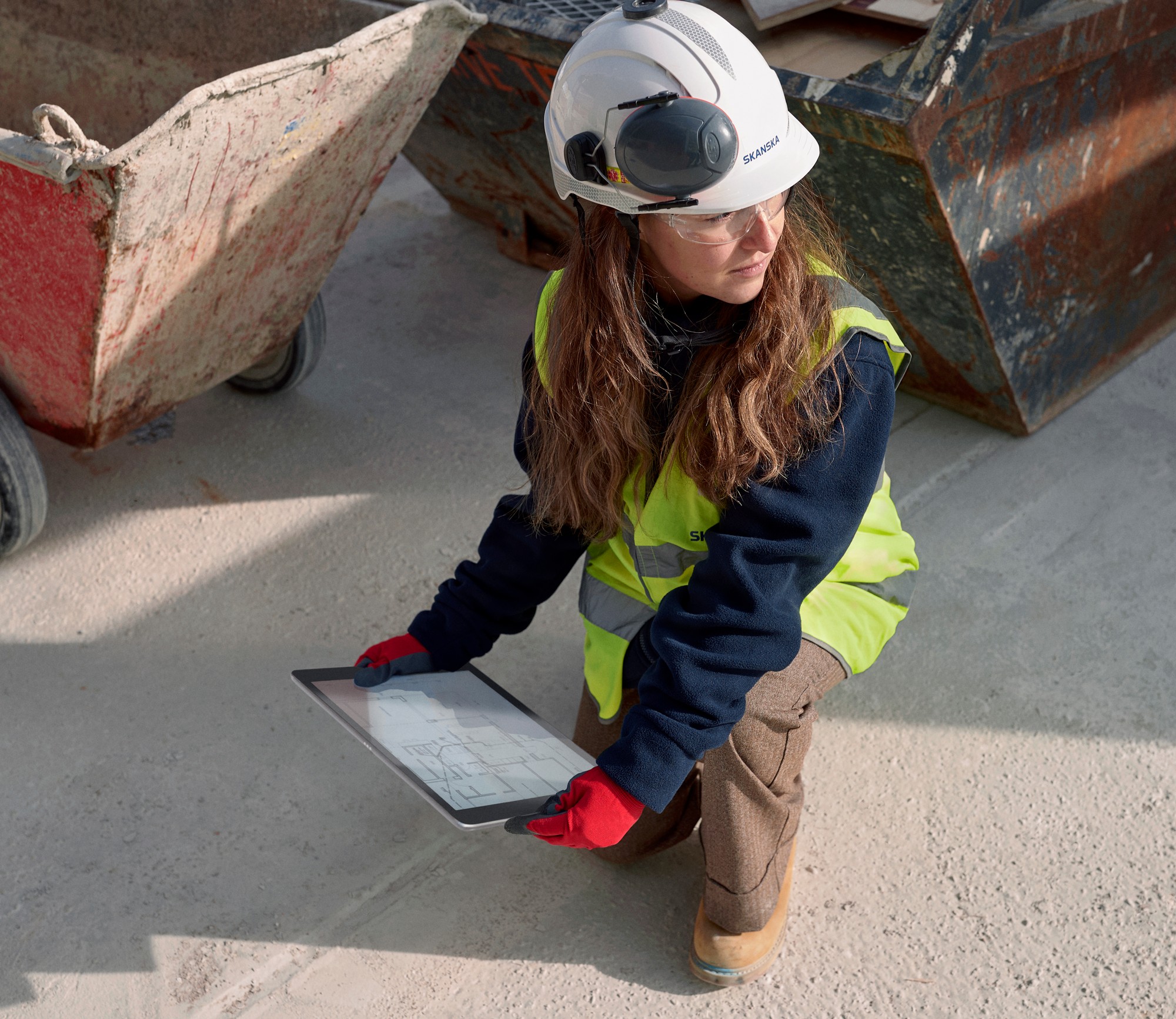 Construction worker in safety gear kneeling with a tablet, examining plans at a job site.