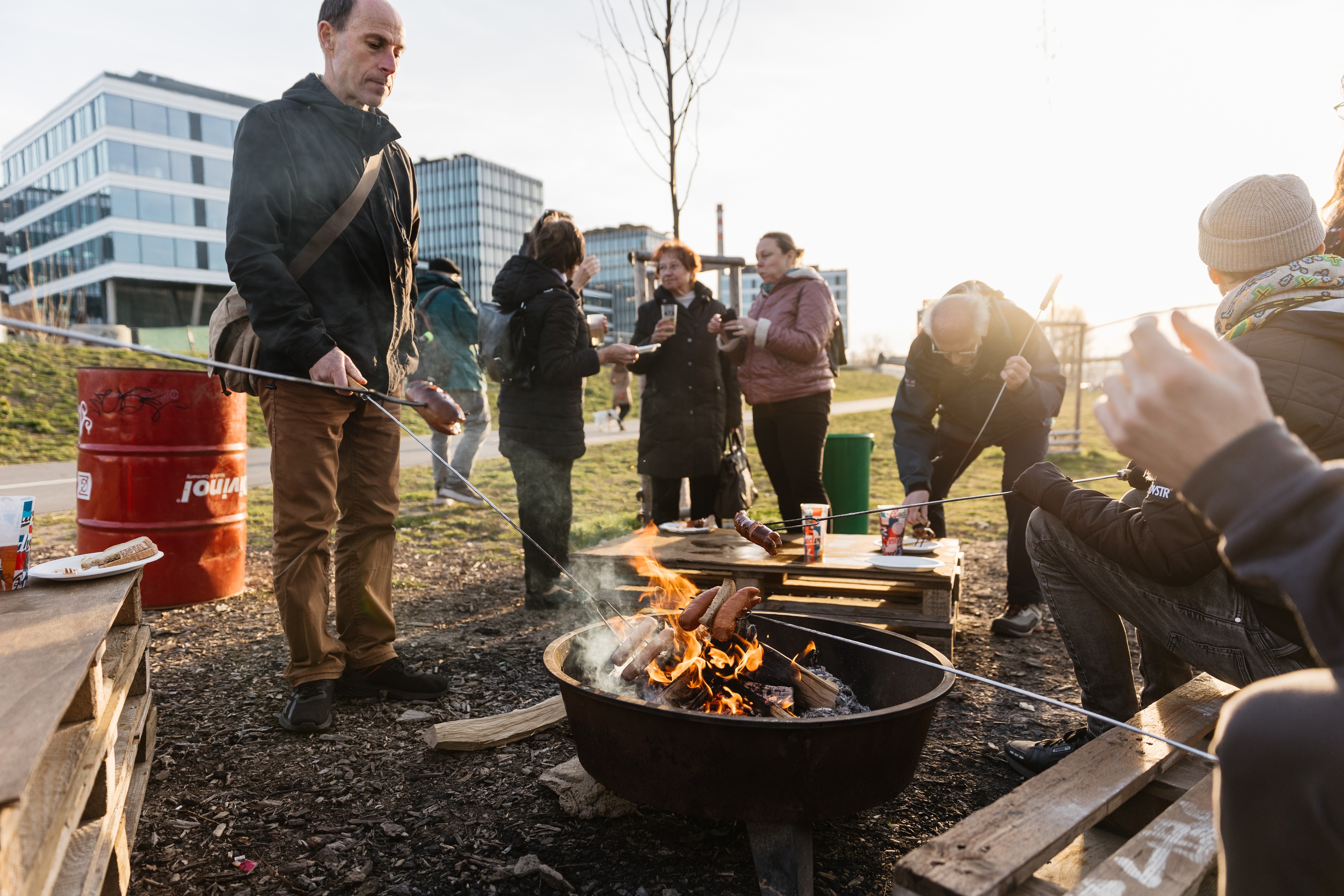 Roasting sausages at Náplavka 2.0 People roasting sausages over an open fire in an urban outdoor environment during the day.