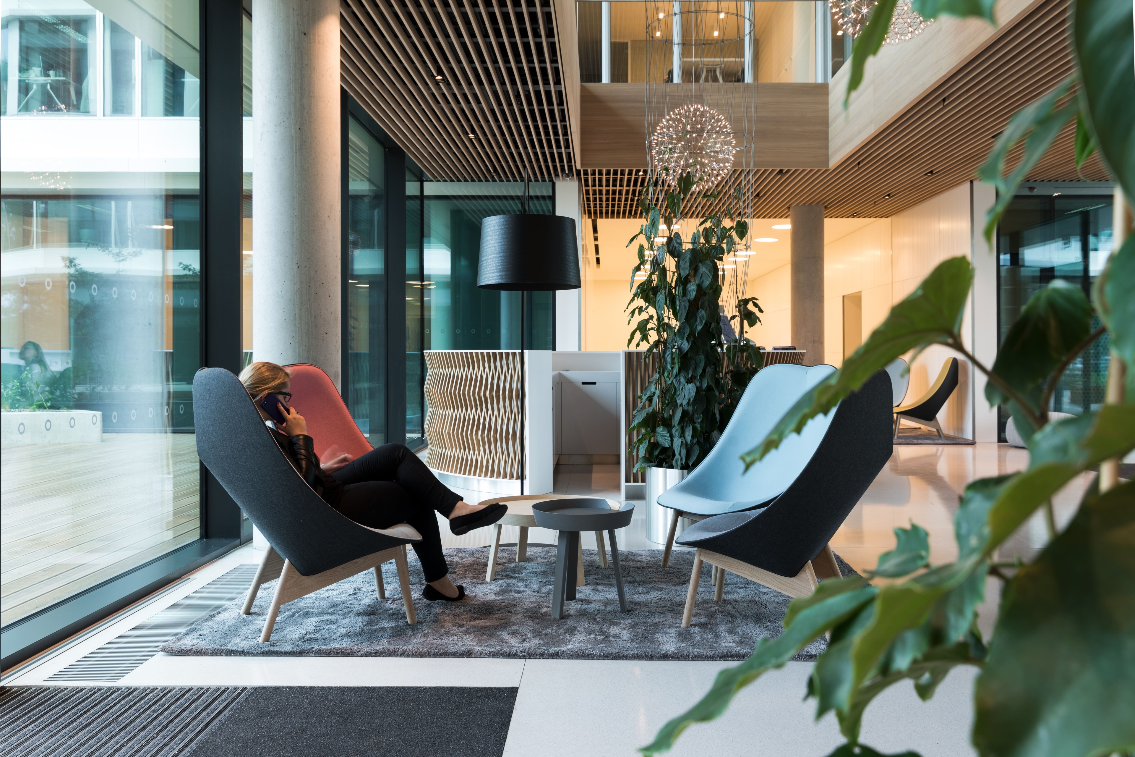 Moderní kancelářská lounge s ženou na telefonu, stylovými židlemi a pokojovými rostlinami.