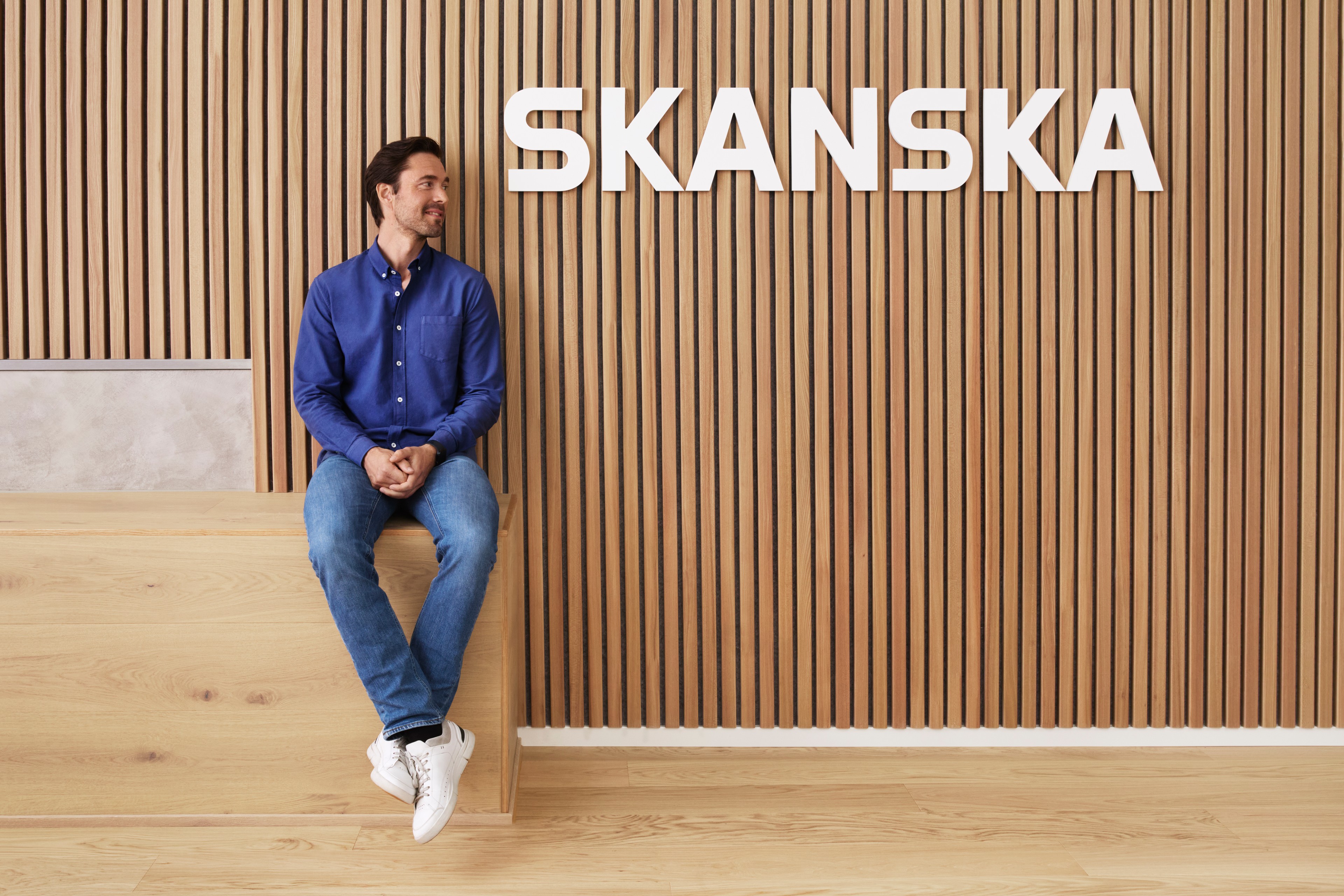 Mężczyzna w niebieskiej koszuli i dżinsach siedzi przy drewnianej ścianie z logo Skanska u góry.