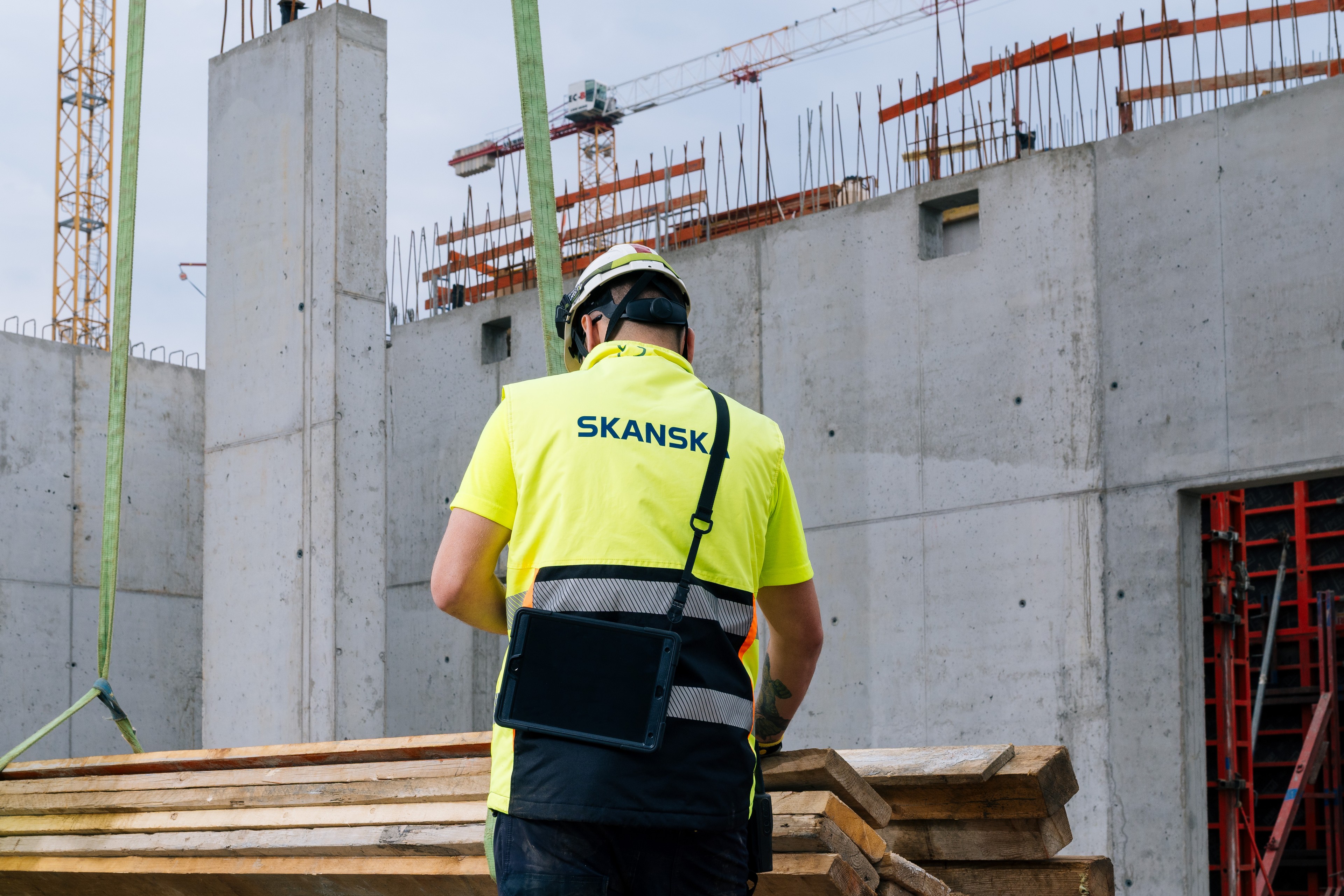 Stavební dělník nosící vestu Skanska na staveništi s betonovými zdmi.