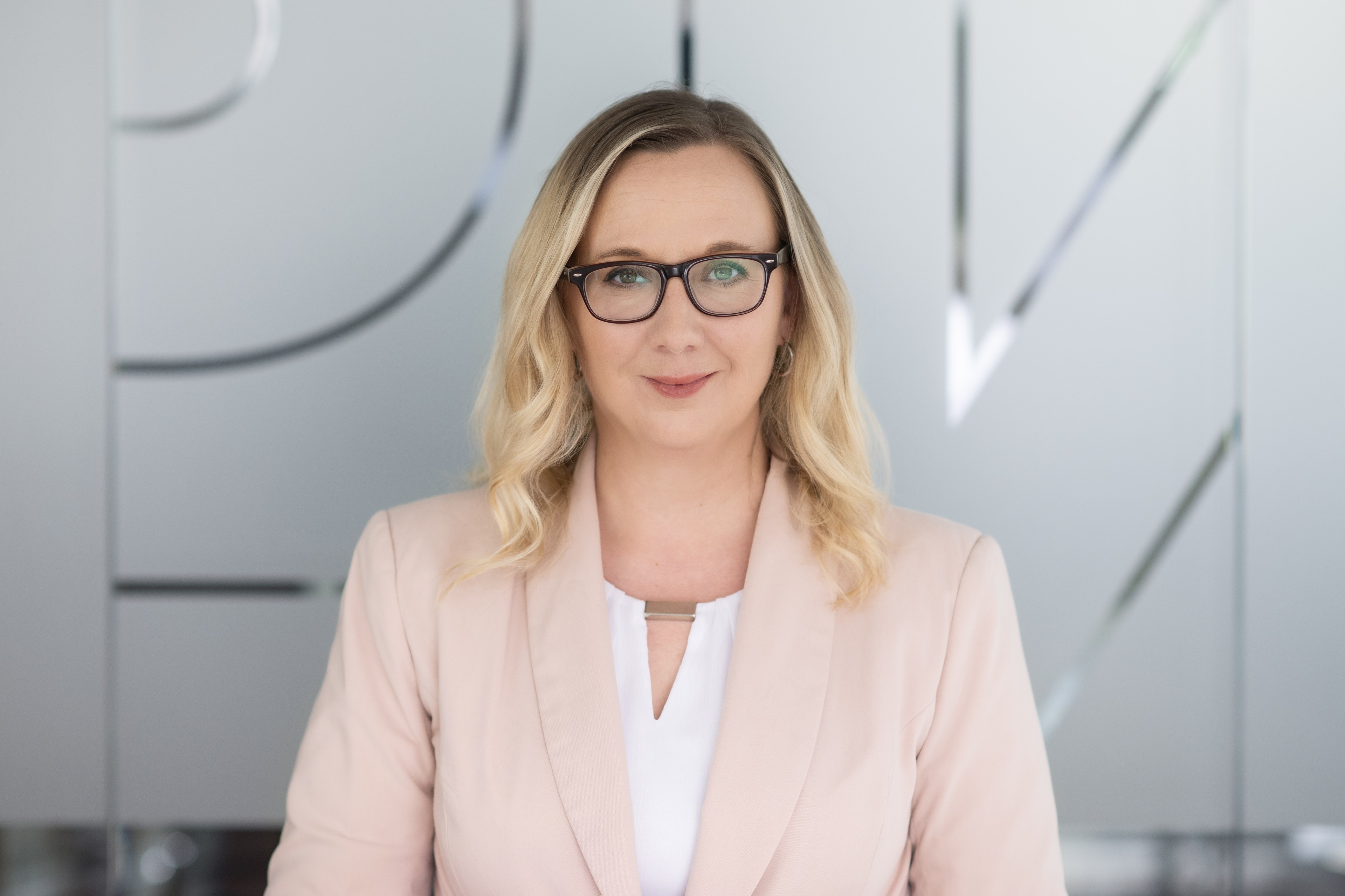 Kateřina Hercogová Profesionální žena v béžovém saku stojící v moderní kanceláři s rozmazanou tváří.