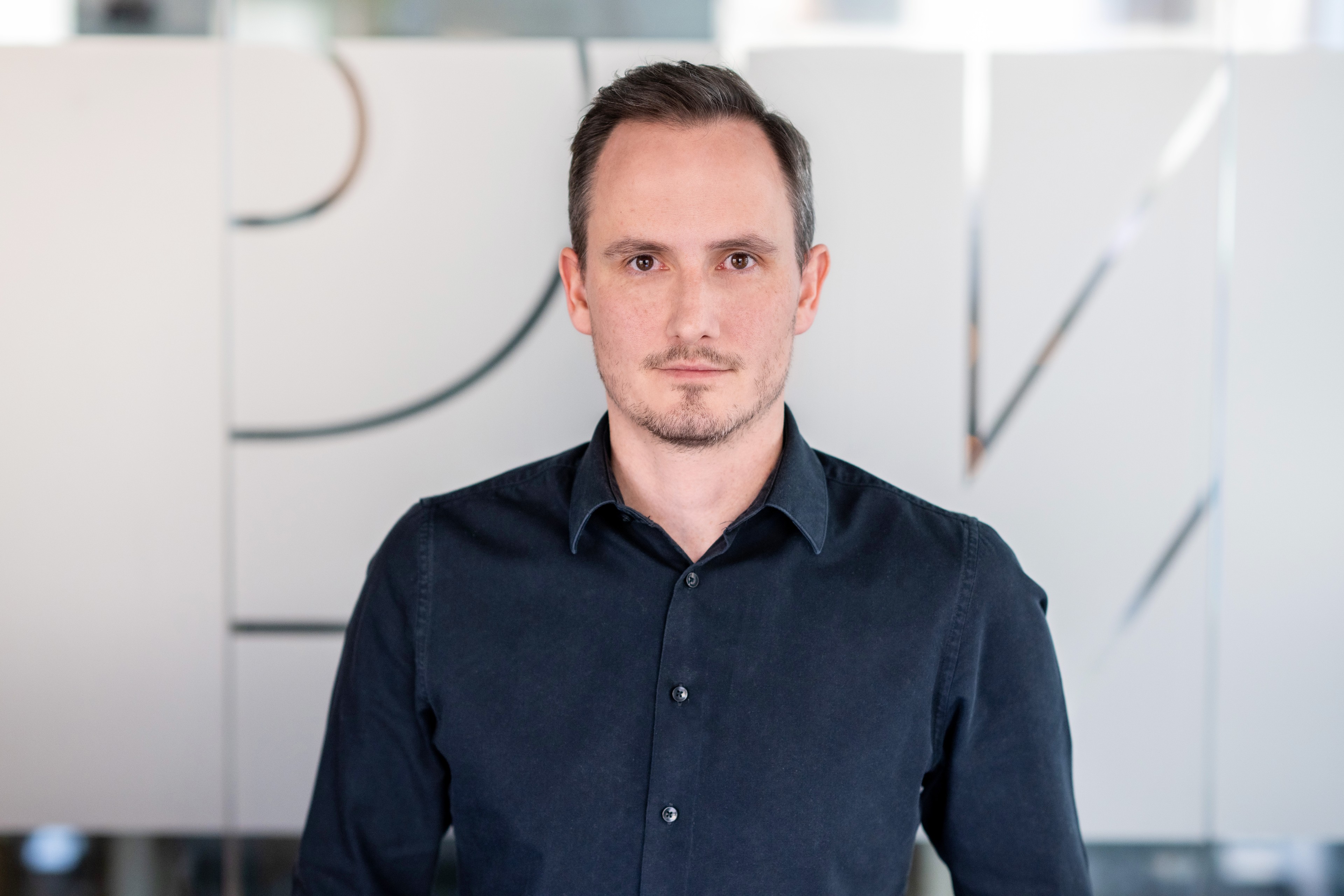 Zbyněk Nývlt Muž v tmavé košili na knoflíky stojící v moderním kancelářském prostředí.