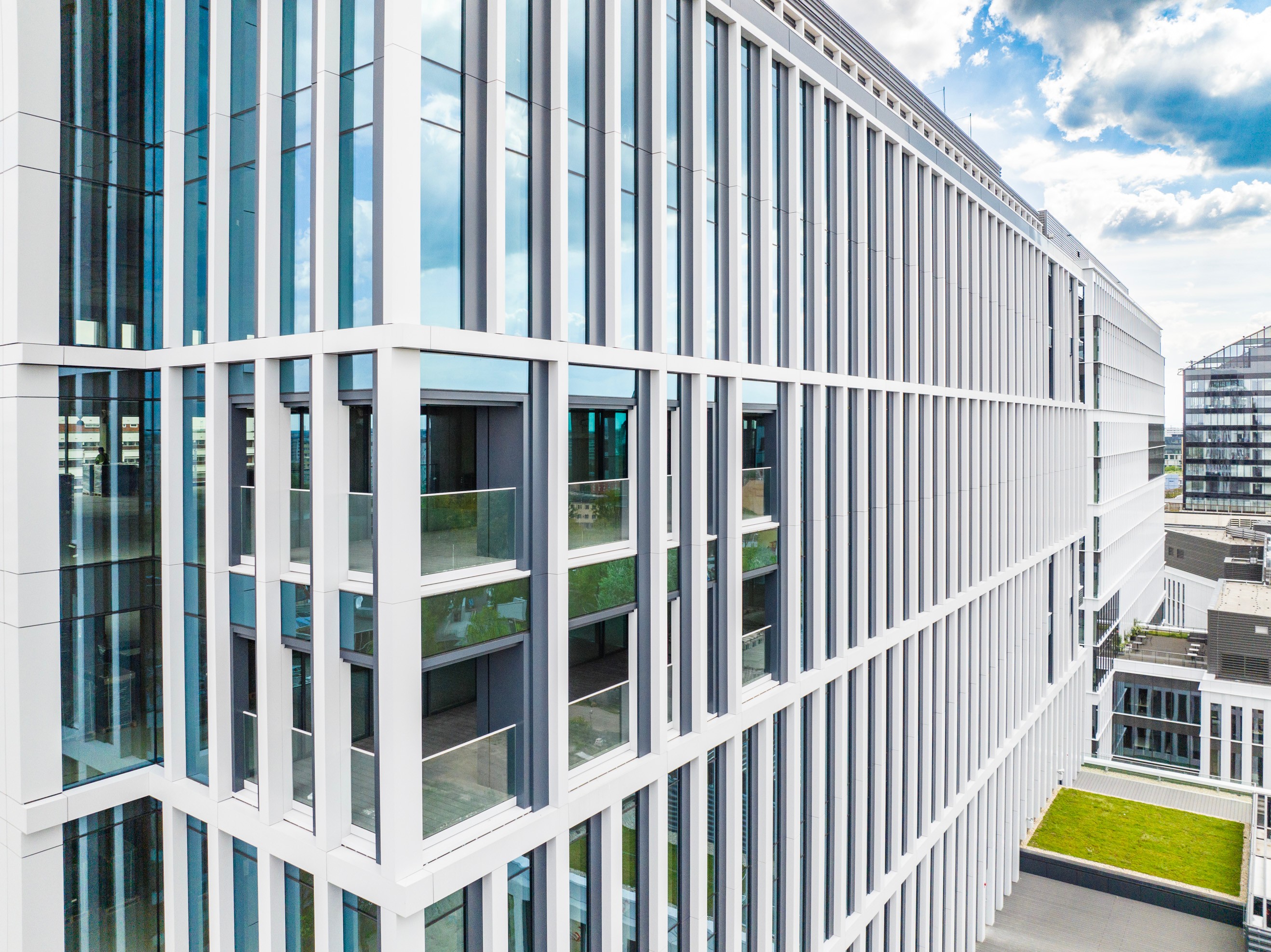 Modern office building with glass facade and vertical white panels under a cloudy blue sky.
