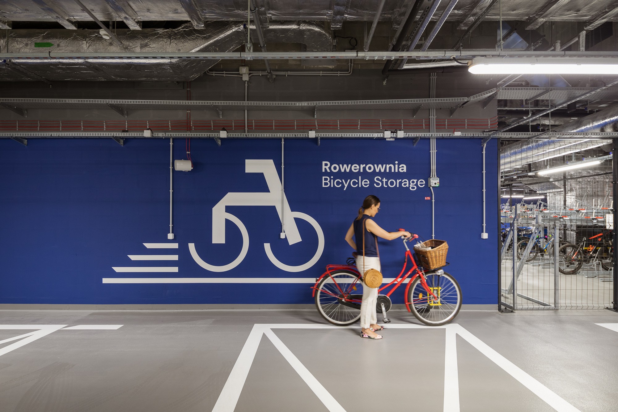 Modern bicycle warehouse with a woman and a red bicycle inside A woman with a red bicycle standing in a modern, indoor bicycle storage area with signage.
