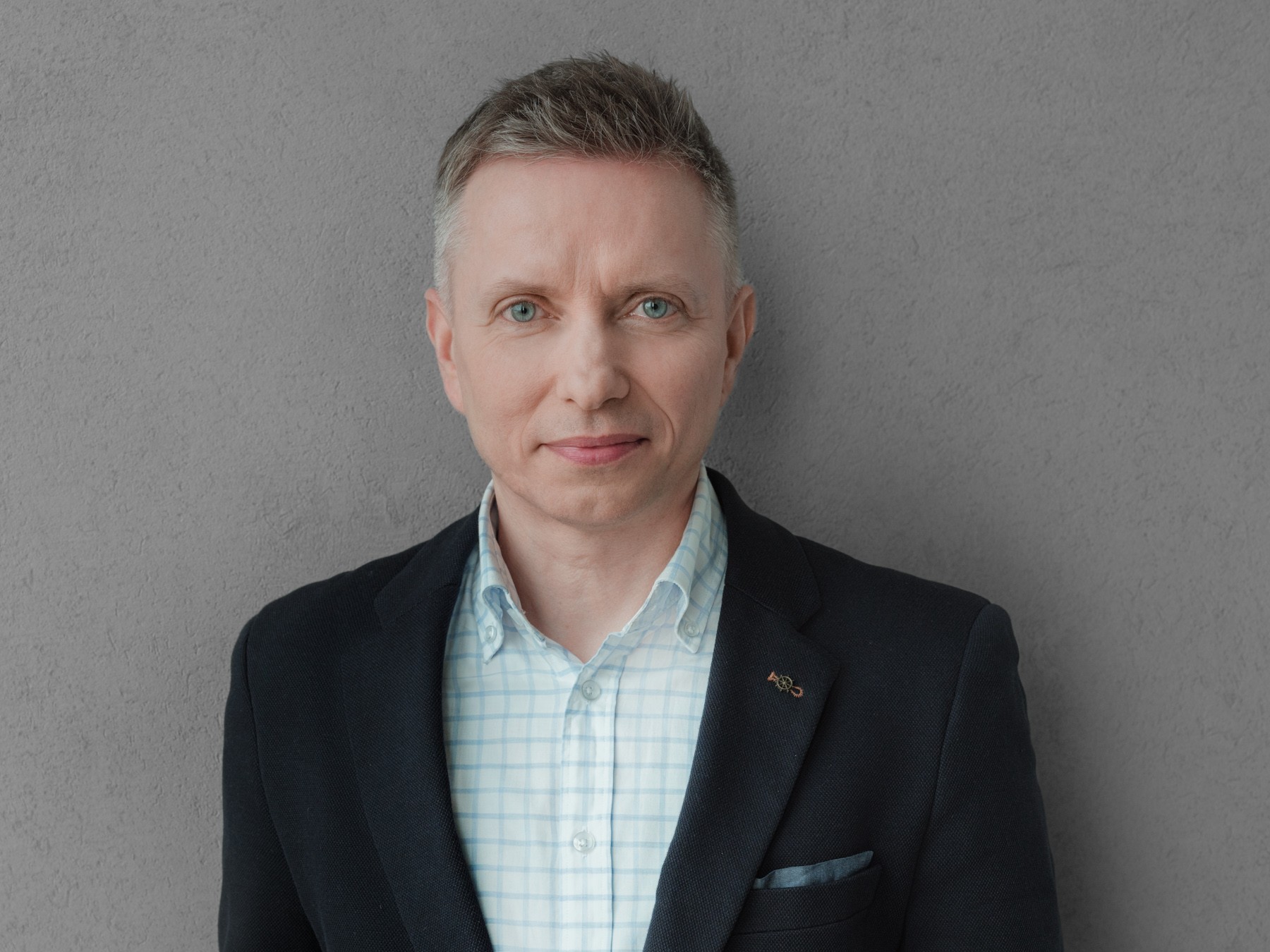 A professional man in a blazer and checkered shirt standing by the wall A professional man dressed in a navy blazer and a checkered shirt against a gray wall.