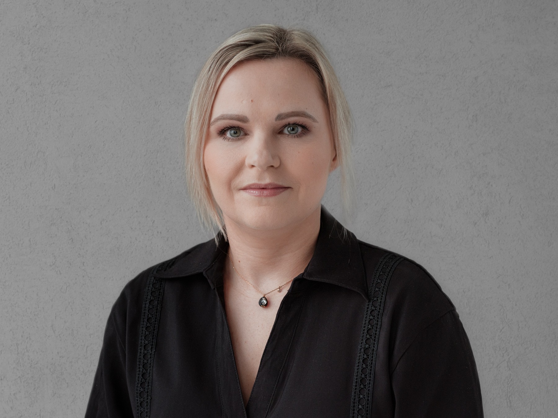 Professional woman in a black shirt with a necklace on a gray background A woman in a black blouse with a necklace with a pendant against a gray textured wall.