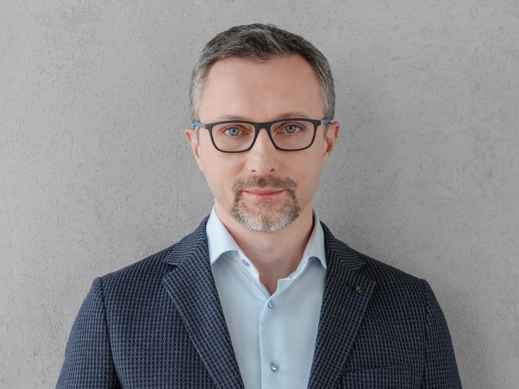 A professional man in a blue blazer and light blue shirt A professional man in a navy blazer and a light blue shirt against a gray wall.