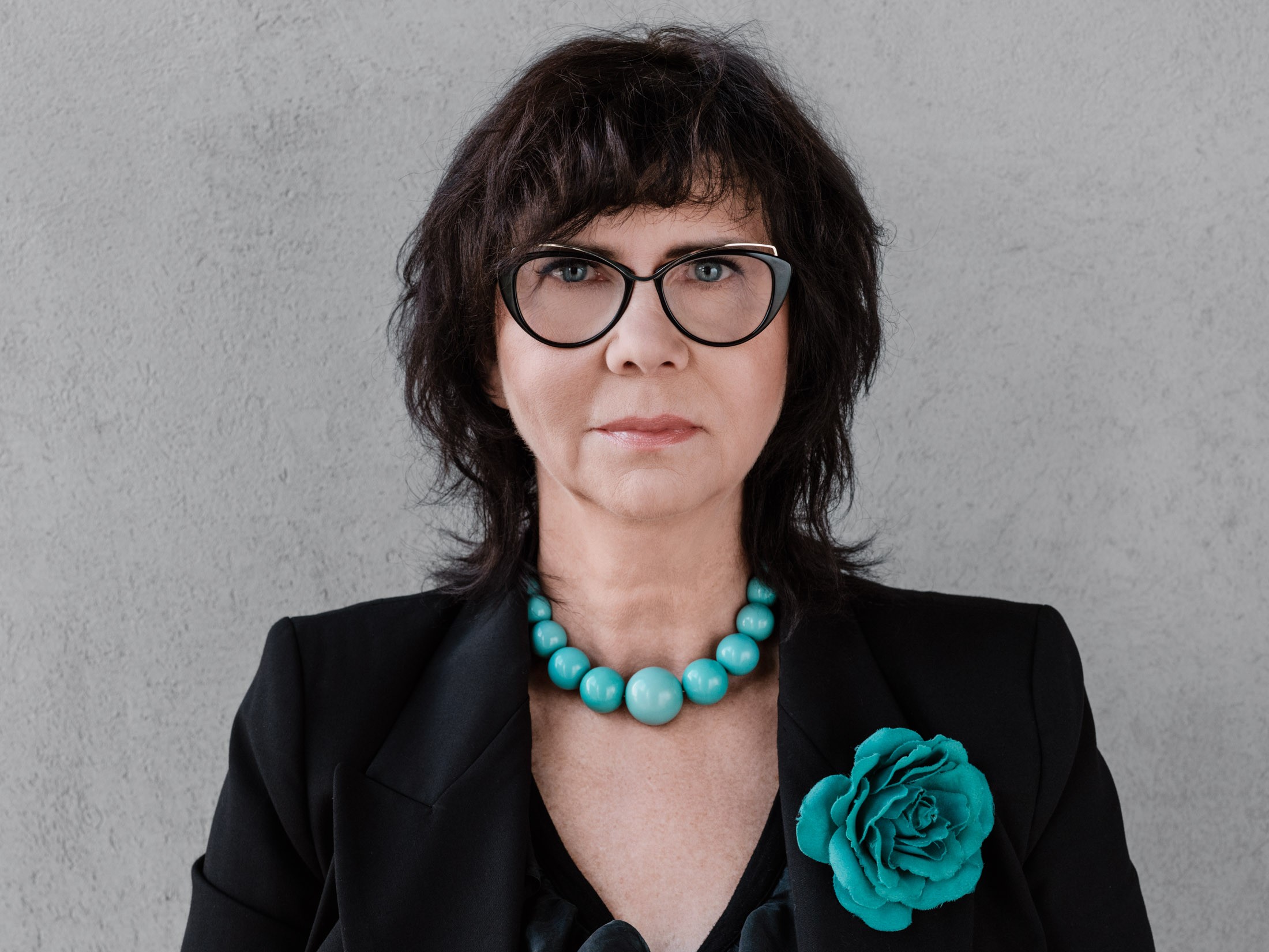 A professional woman wearing a turquoise necklace and a flower-shaped brooch A woman in a black blazer with a turquoise necklace and a flower-shaped brooch against a gray wall.