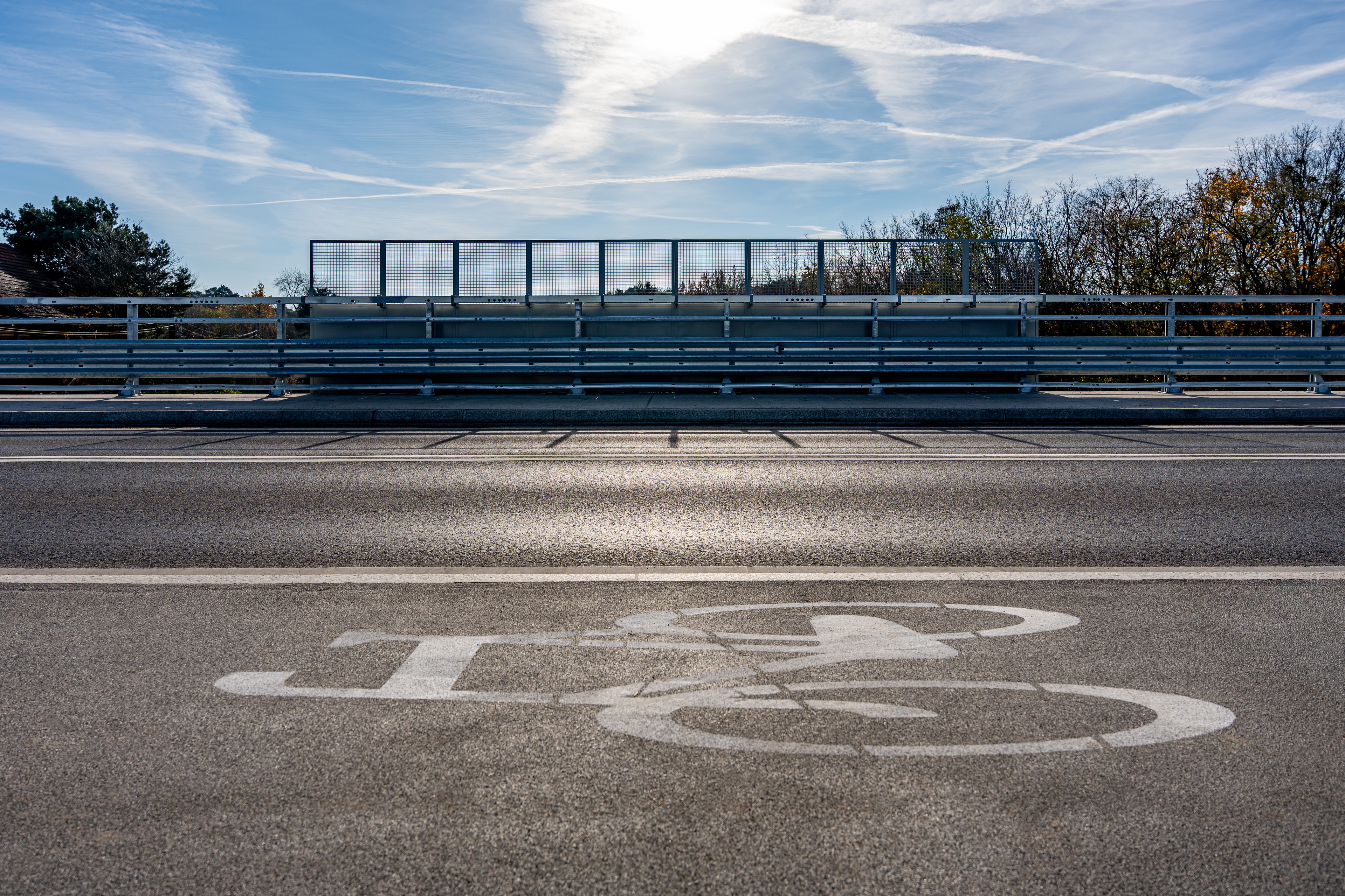 Prázdná silnice s vyznačenými jízdními pruhy pro cyklisty a kovovou zábranou pod jasnou, částečně oblačnou oblohou.