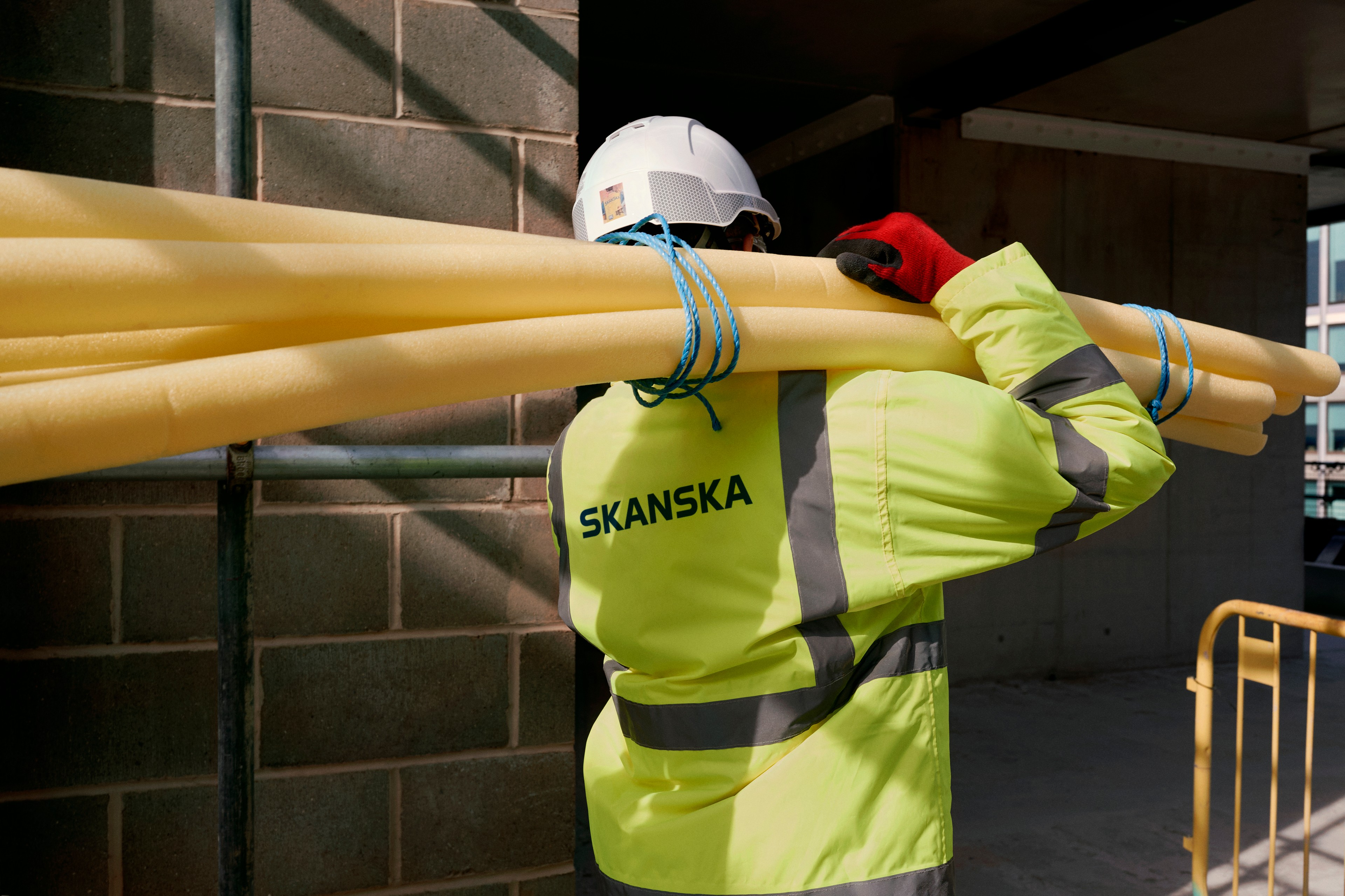Construction worker carrying yellow foam pipes, wearing a safety vest and helmet.