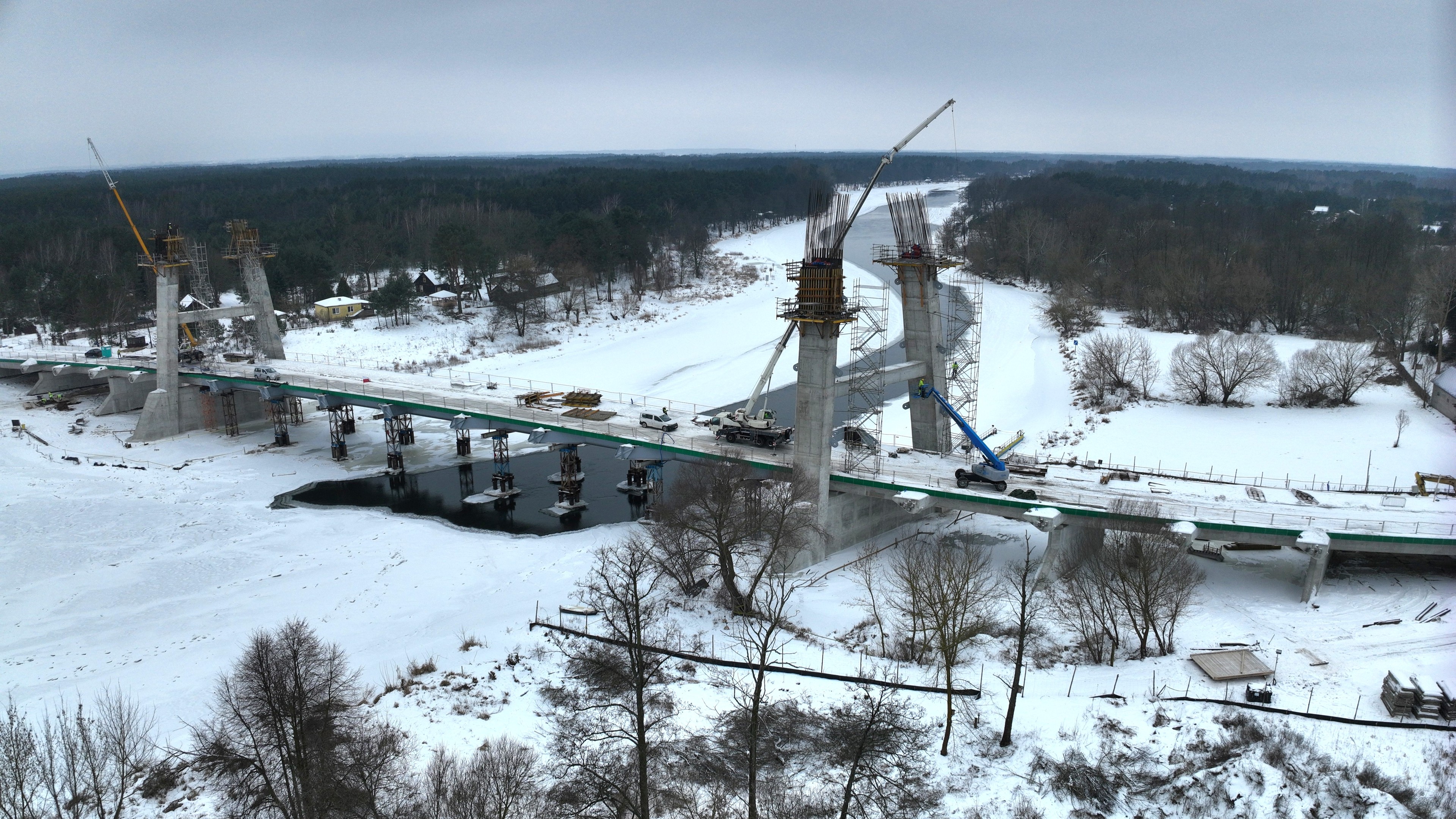 Snowy Bridge Construction Over River in Winter Landscape Most pokryty śniegiem w trakcie budowy nad zamarzniętą rzeką, z widocznymi dźwigami i rusztowaniami.