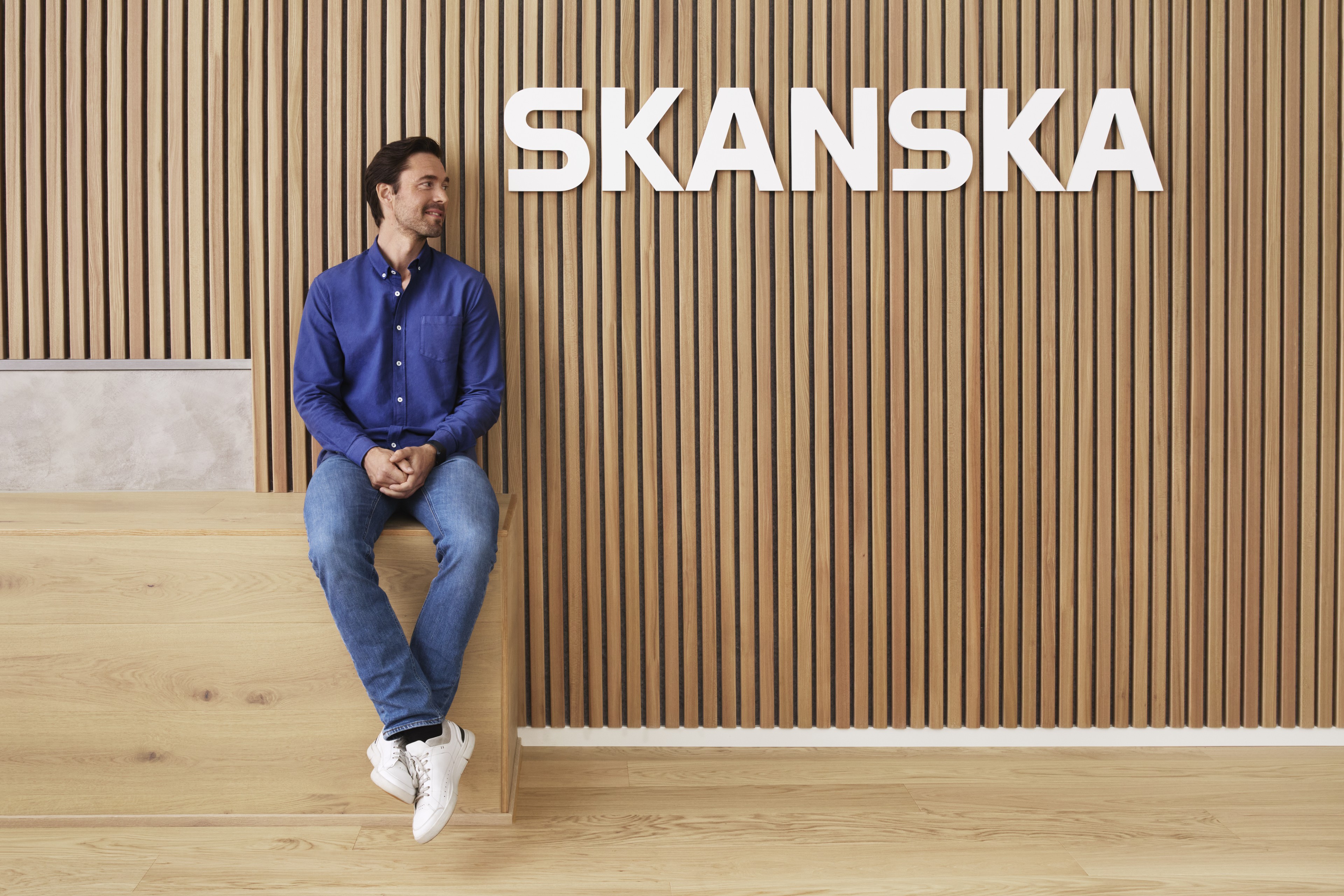 A man in a blue shirt and jeans is sitting by a wooden wall with the Skanska logo above.
