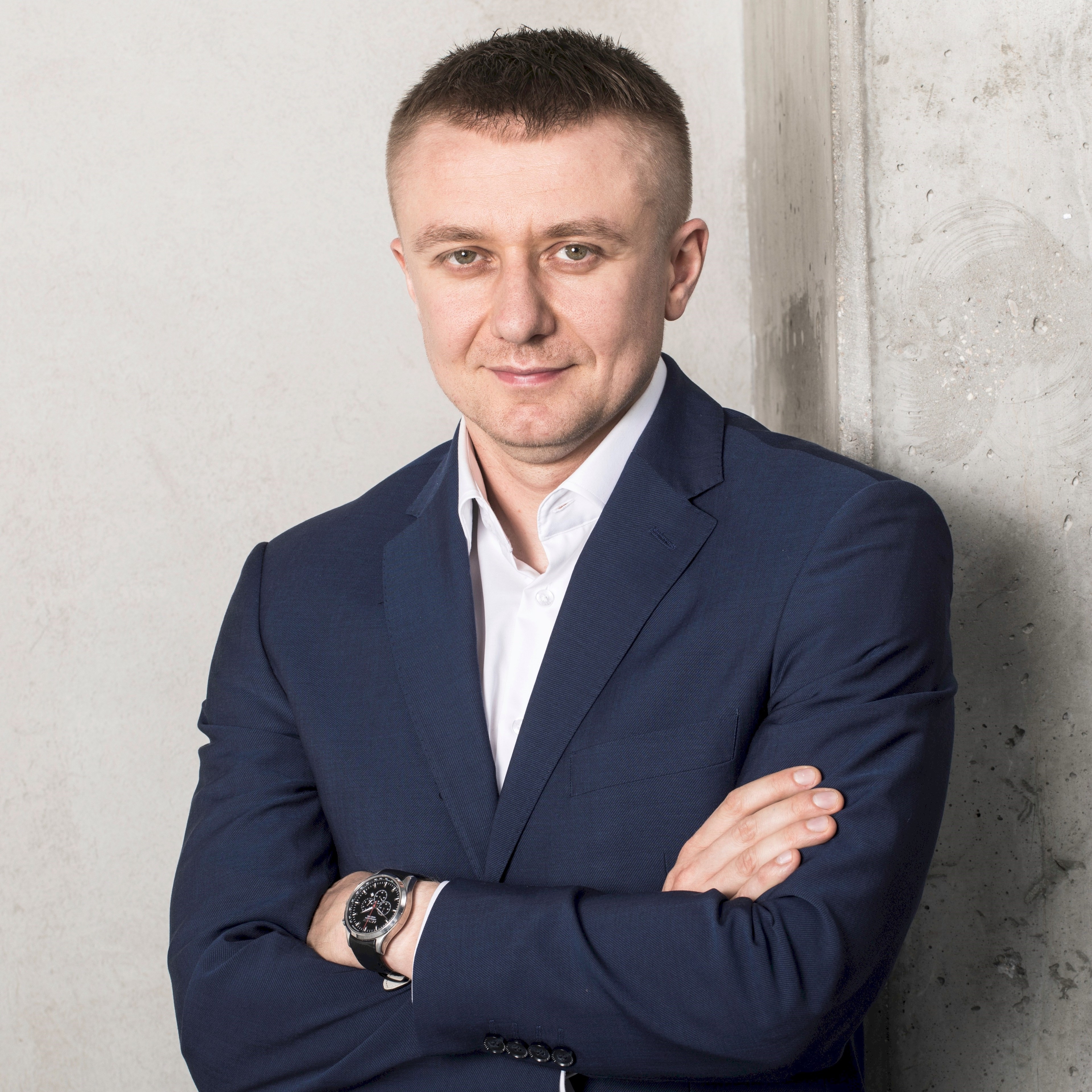 A man in a navy blue suit with crossed arms, wearing a watch on his wrist, standing against a concrete wall.