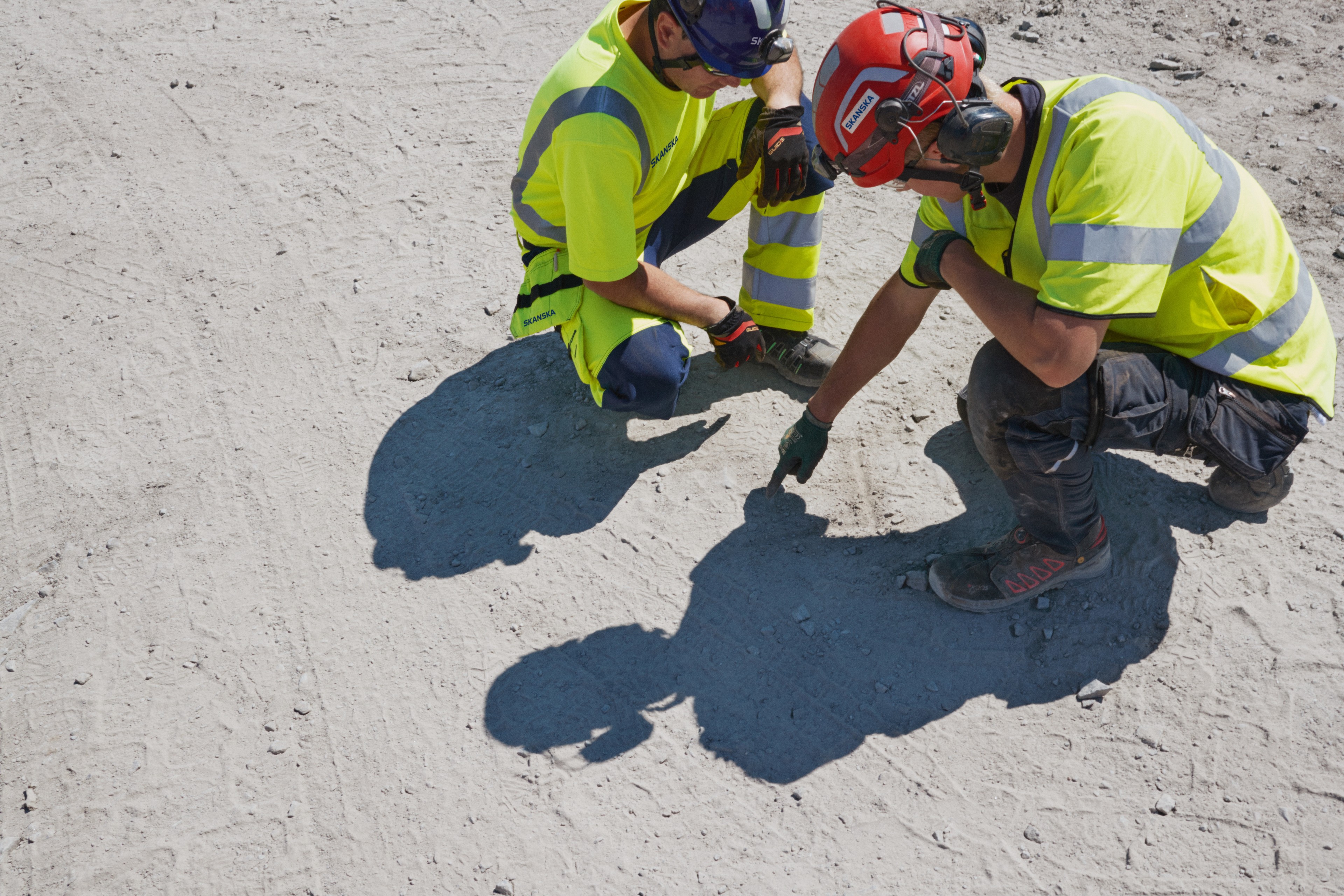 Two construction workers in protective clothing examining the soil on a dusty site.