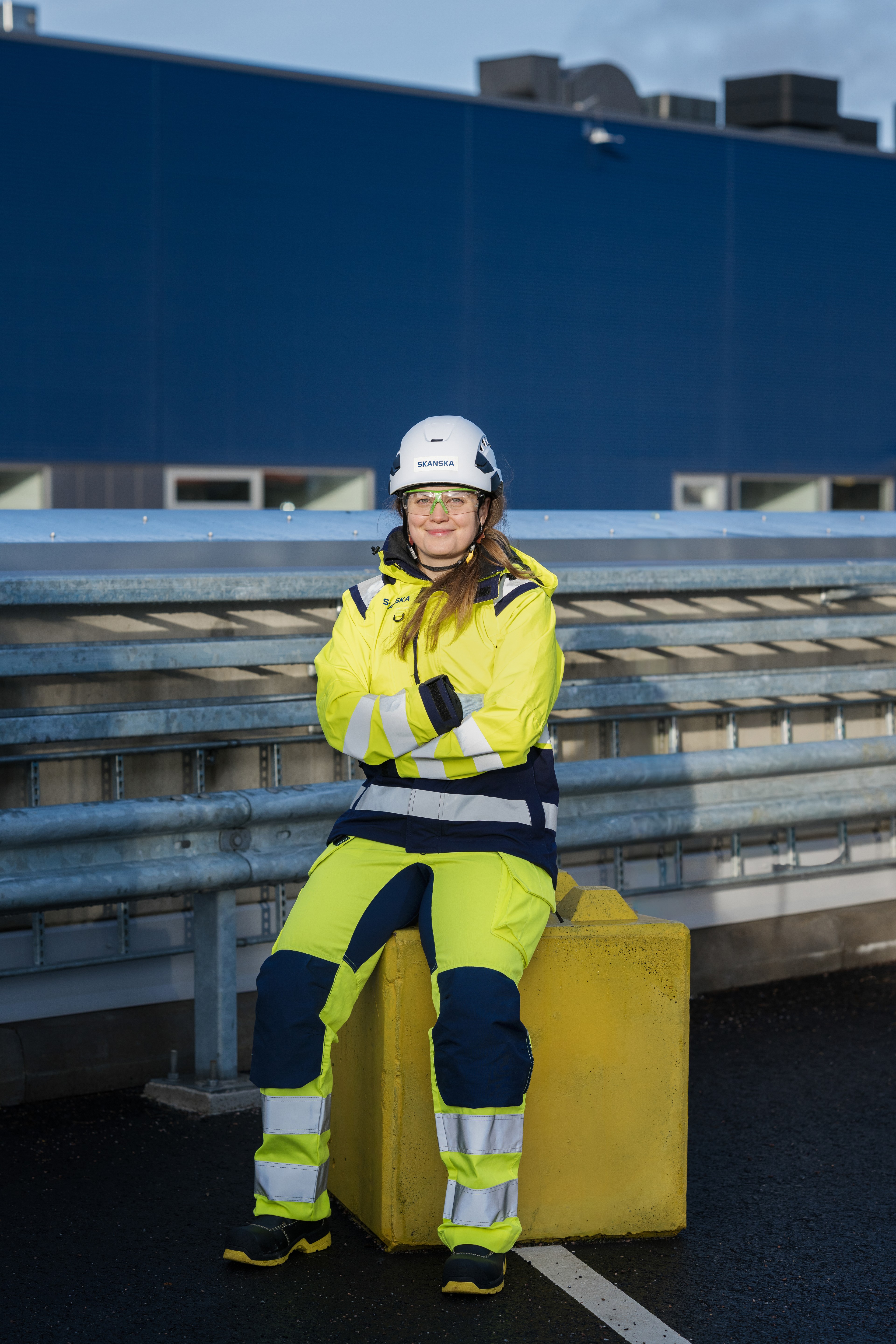 A person in high-visibility protective clothing and a helmet sitting on a yellow block outside.