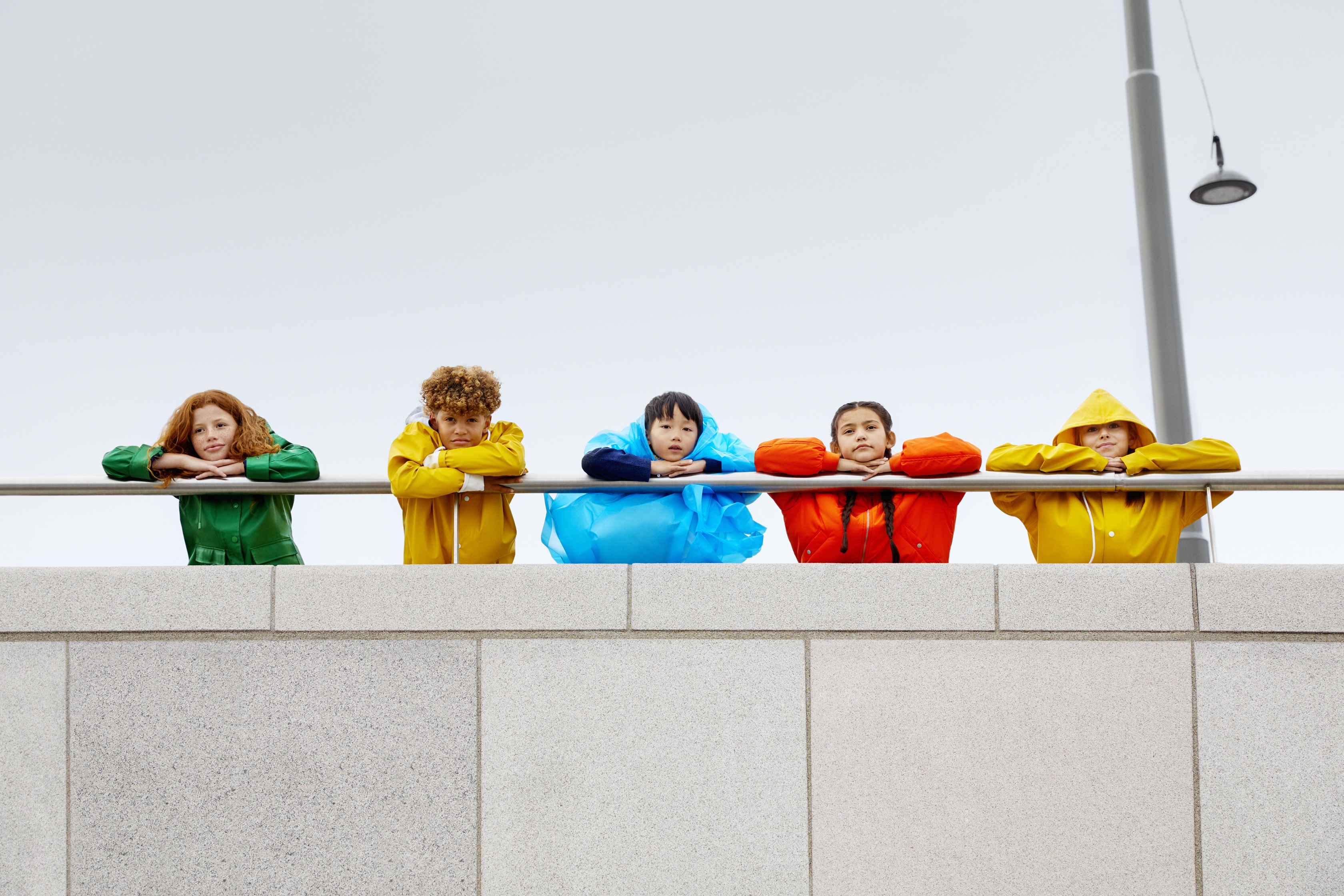 Five people in colorful raincoats leaning on a railing against a gray sky.