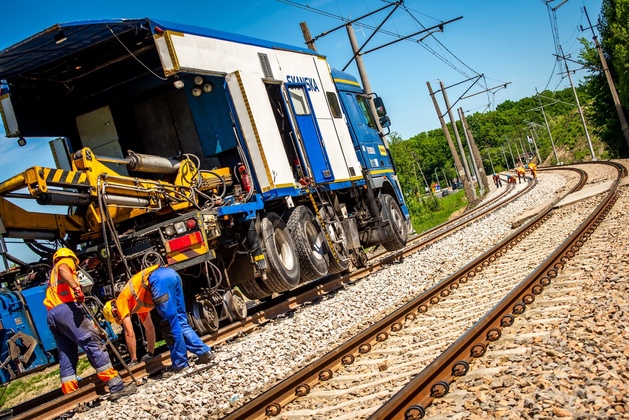 Railway Maintenance Team Working on Tracks Robotnicy naprawiający tory kolejowe dużym pojazdem konserwacyjnym pod czystym, błękitnym niebem.