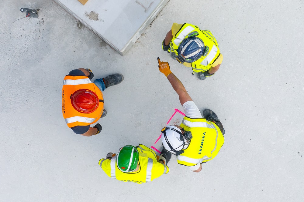 Stavební dělníci v ochranném vybavení probírají plány na betonovém prostranství, viděni shora.