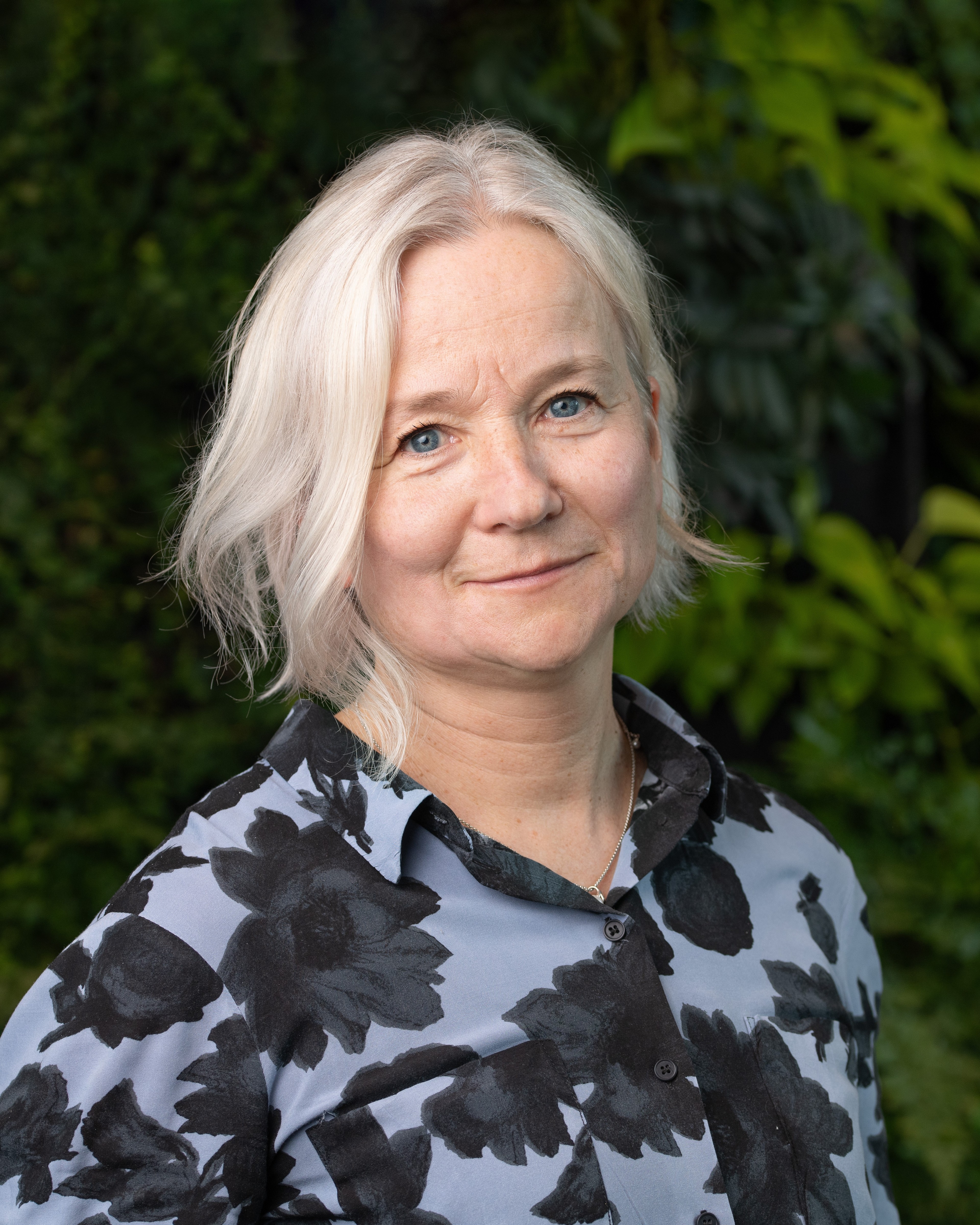 Camilla Wieslander Kvinna som bär en svartvit blommig skjorta med grönskande bakgrund.