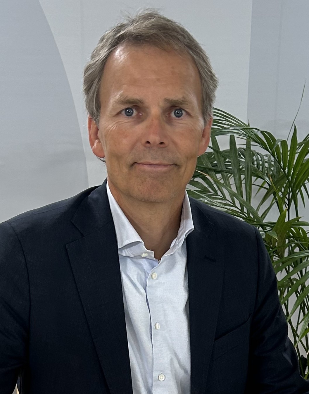 Jan Odelstam Man i en mörk kostym och vit skjorta som sitter inomhus med gröna växter i bakgrunden.