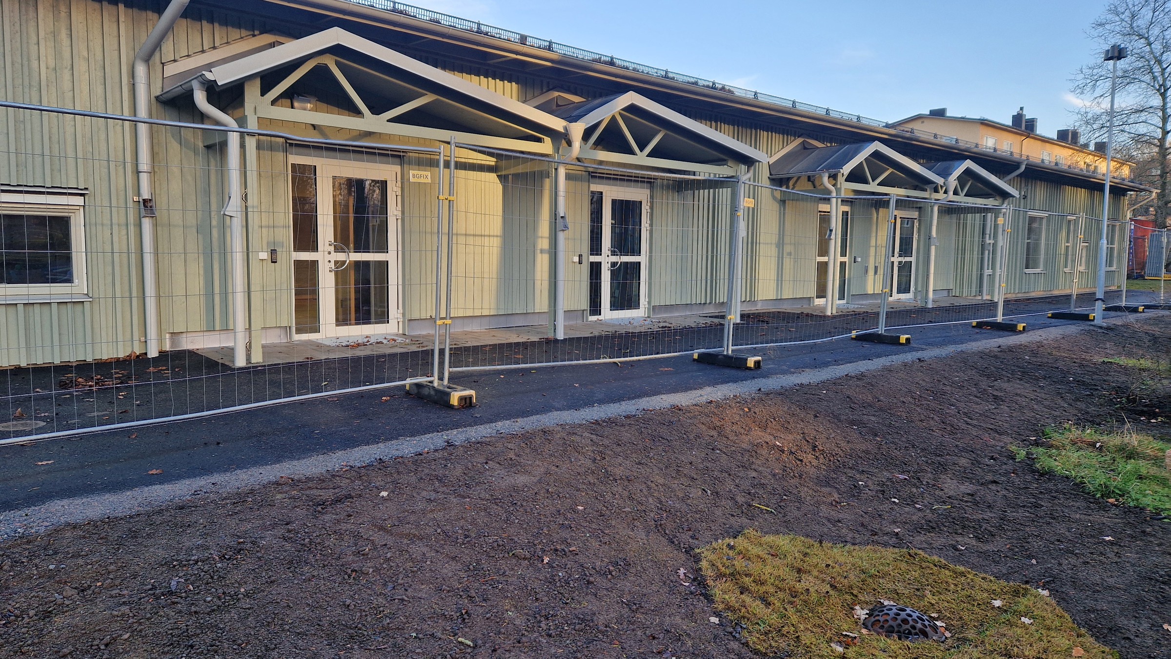 Bägersta gård förskola har en grön stående träpanel och ett mörkgrått plåttak som harmonierar fint med den naturliga omgivningen. Bägersta gård förskola med ett byggstaket i förgrunden.