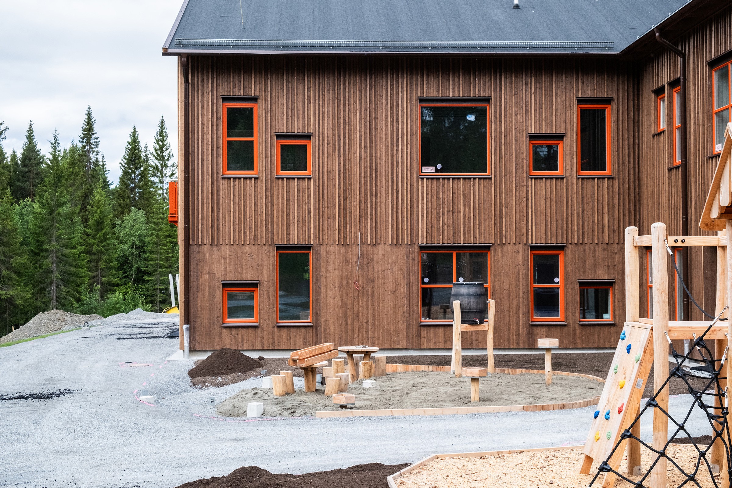 Fasad i brunt trä där två huskroppar möts i en hörna. Framför syns lekredskap under uppbyggnad.