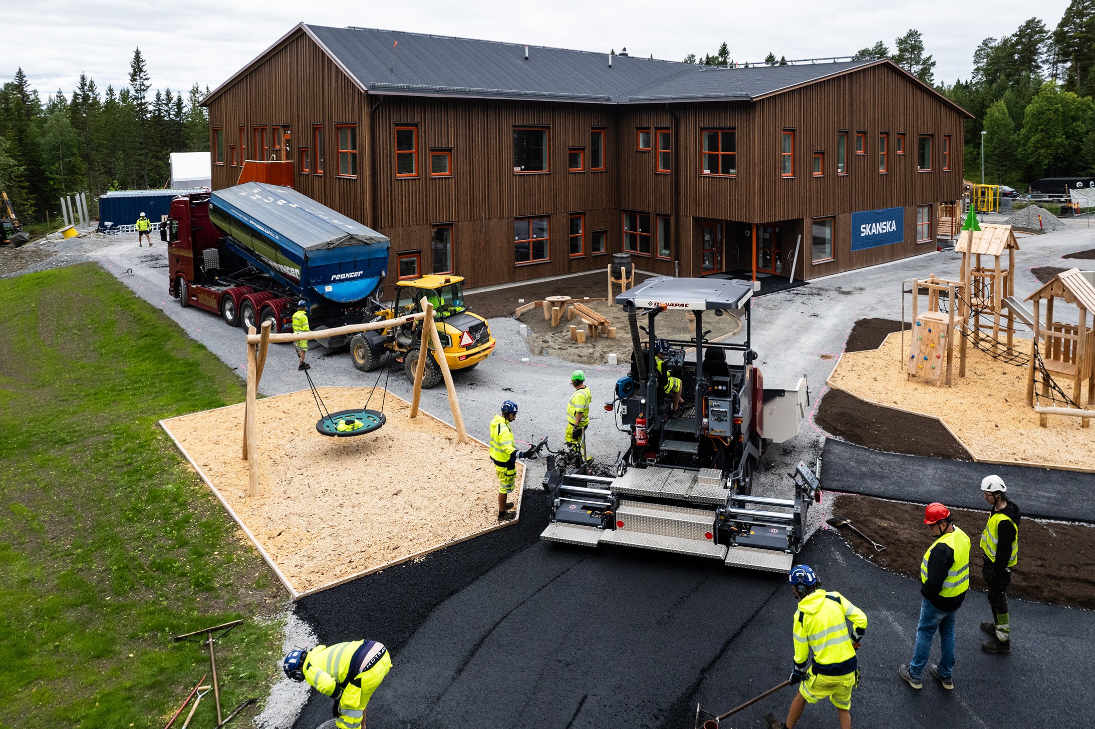 Asfaltläggare i arbete på skolgård med personer i gula varselkläder intill.