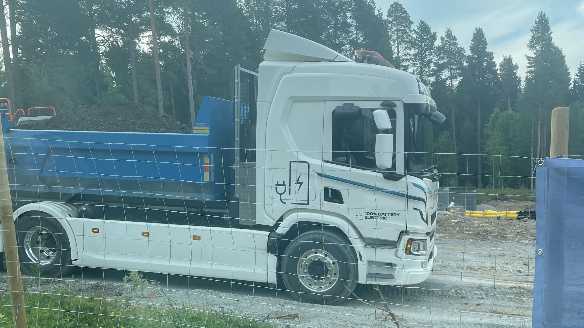 Elektrisk dumper parkerad på en byggarbetsplats, omgiven av träd och inhägnad av ett staket.