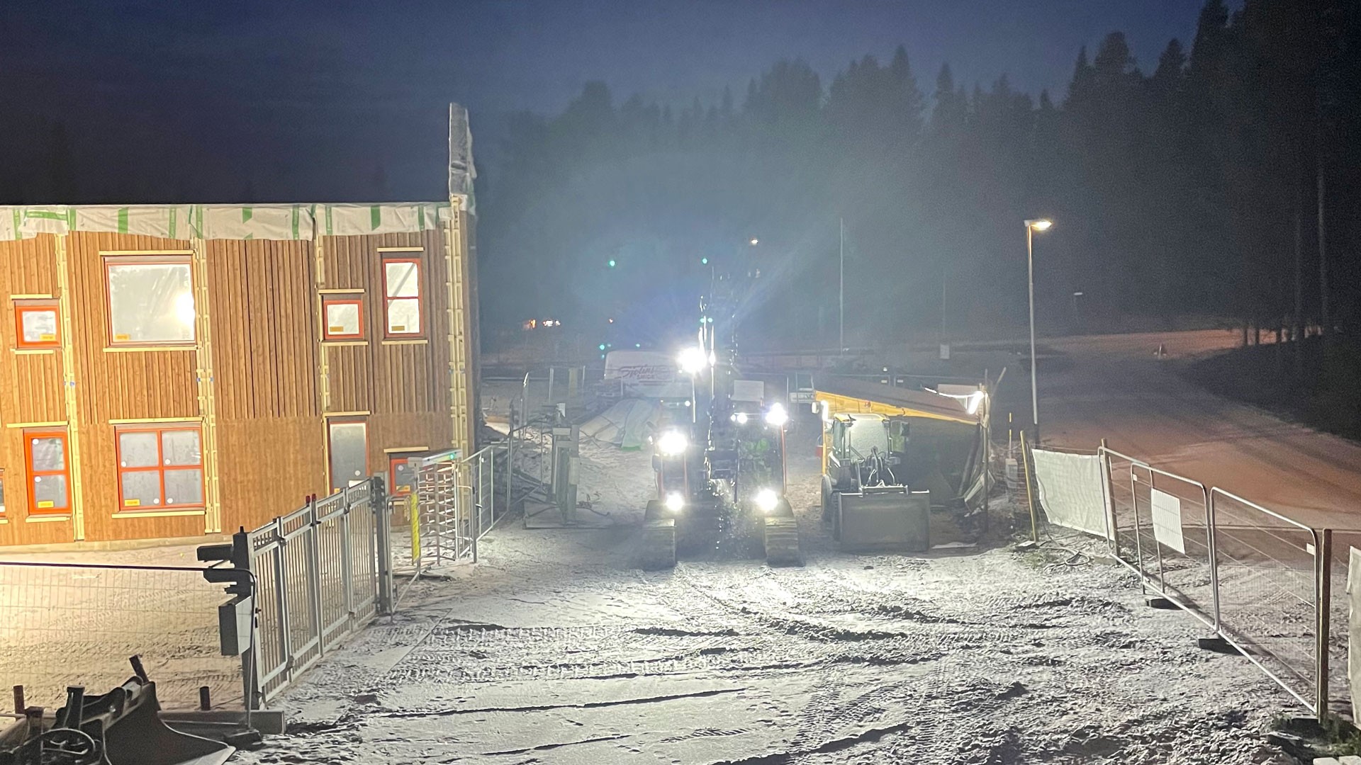 Nattlig byggscen med maskiner och en delvis byggd trästruktur, omgiven av snö.