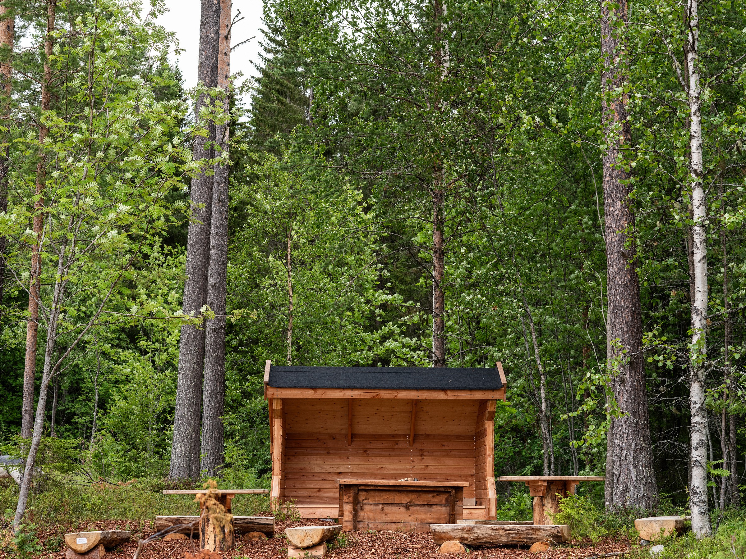 Trähus med öppen framsida, intill finns träbord och bänkar av stockar.