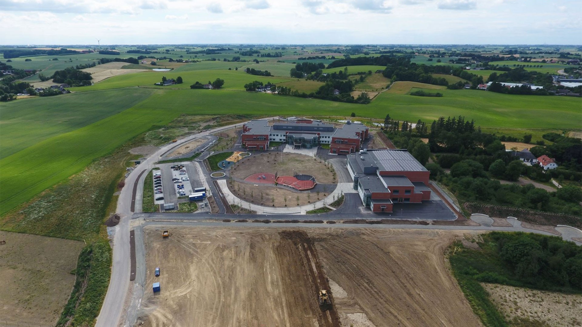 Drönarbild med skolan, rastgård och sporthall i centrum. Åkermark omgärdar skolområdet.