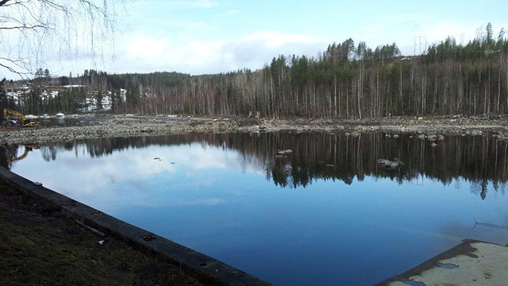 Älvfåran nedströms dammen fick torrläggas två gånger på grund av höstfloden 2011. En lugn sjö som speglar den omgivande skogen och himlen, omgiven av stenar och gles vegetation.
