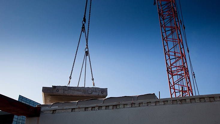 I projektet har prefabricerade stommar använts. En kran lyfter en betongplatta ovanför en byggarbetsplats mot en klarblå himmel.