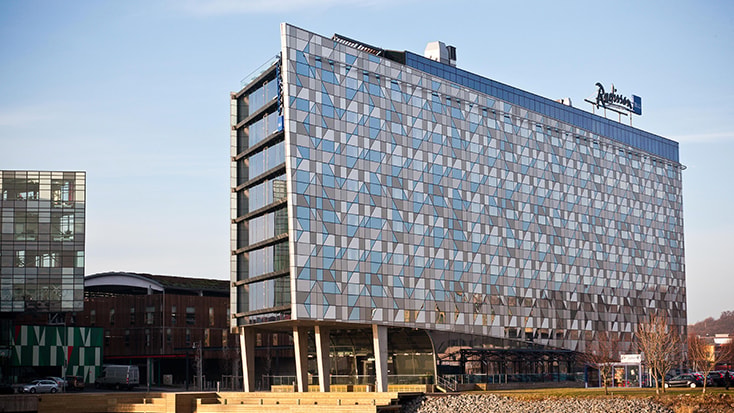 Radison Blu Riverside Hotel. Modern glasfasadhotellbyggnad med Radisson Blu-logotyp, belägen i ett urbant område.