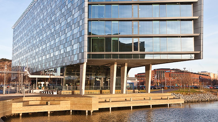 Radisson Blu Riverside Hotel. Modern glasbyggnad på pålar vid en strandlinje, som reflekterar solljus, med urbana omgivningar.