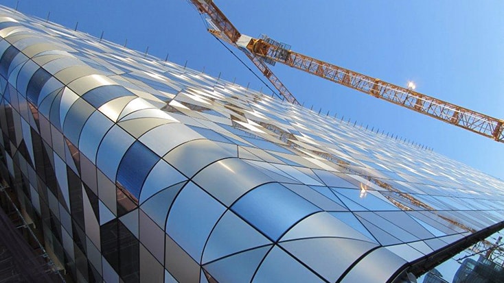 Till fasaden användes färdiga ytterväggselement i glas och aluminium. Modern glasbyggnad under konstruktion med en kran mot en klarblå himmel.
