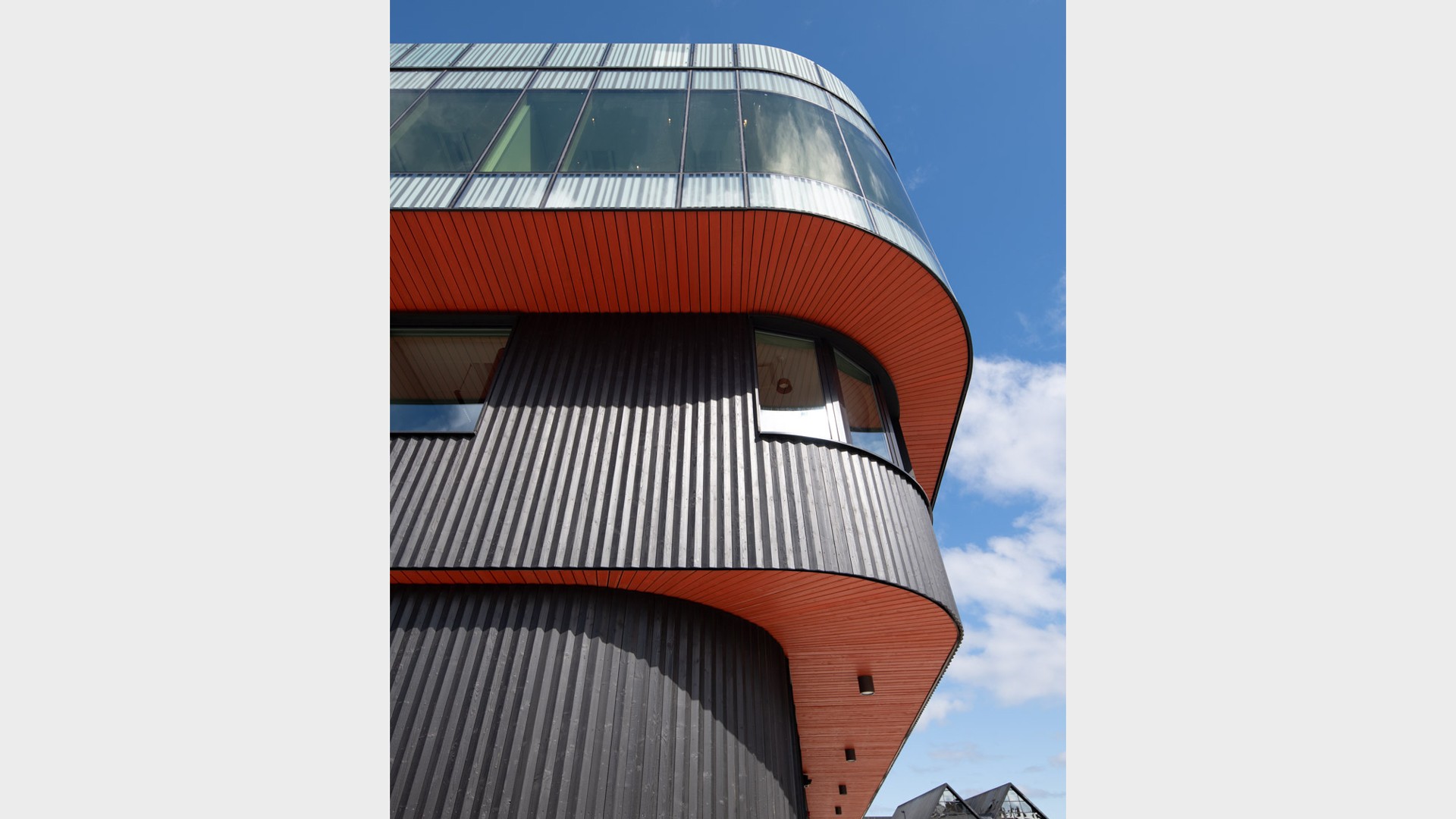 De röda utskjutande undersidorna påminner om den svarttjärade Blekingeekan med röda inslag på botten och durk. Foto: Anders Bobert Närbild av en Boverkets huvudkontor med svarta träribbor och rundad glasfasad under blå himmel.