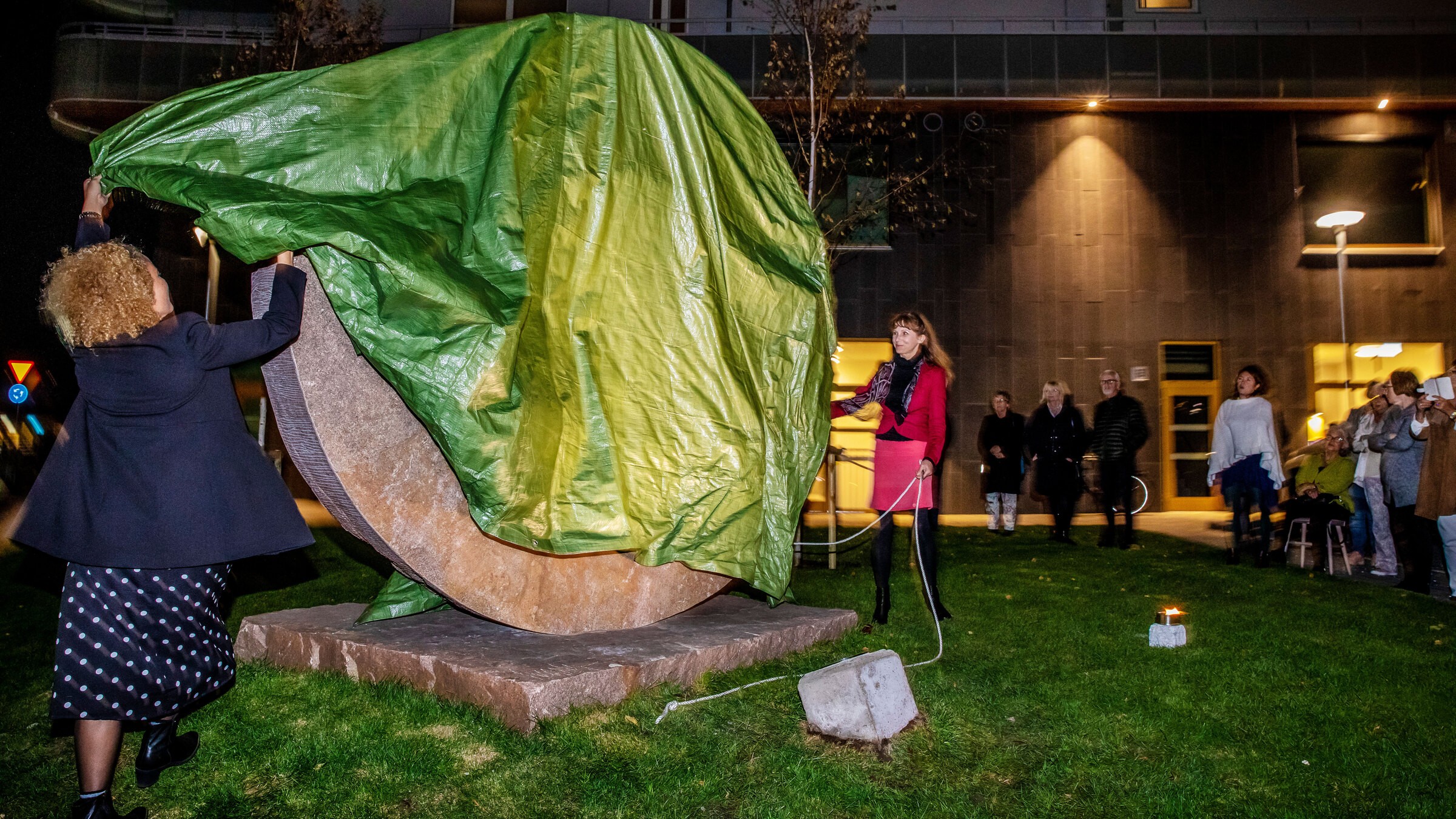 Två kvinnor avtäcker en stor, täckt skulptur utomhus, omgivna av en liten folkmassa på natten.