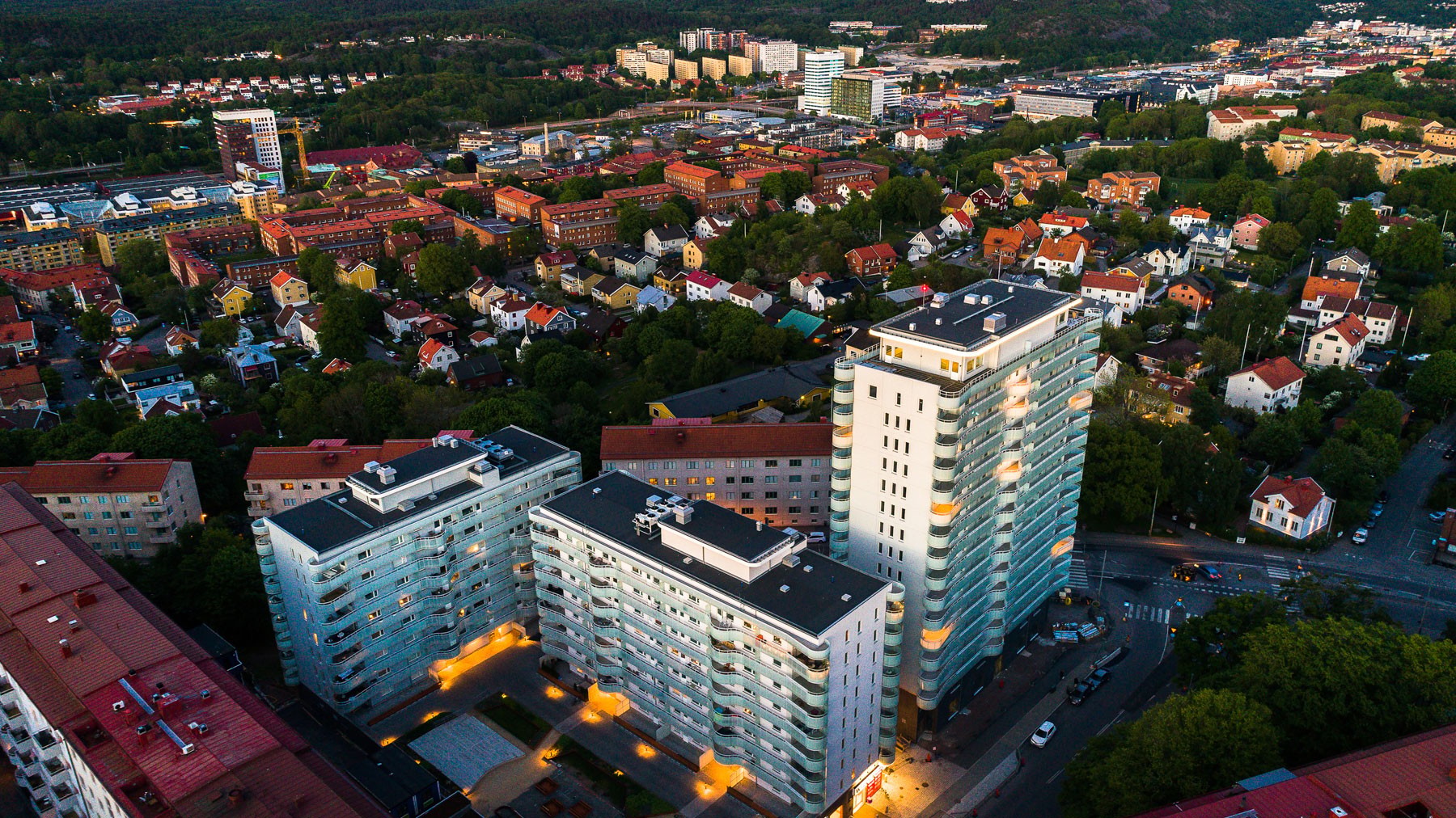 Flygvy av urbana lägenhetsbyggnader med omgivande grönska och stadsbild i skymningen.