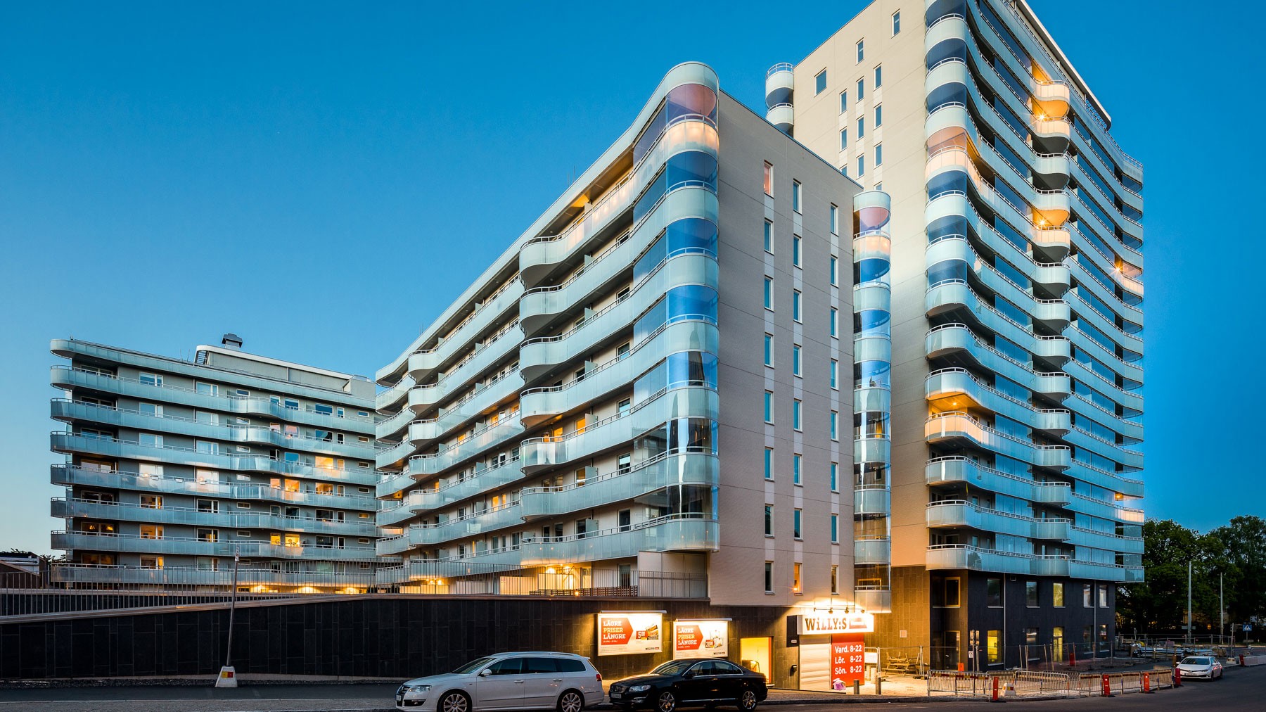 Modernt flerfamiljshus med upplysta balkonger och butiksutrymmen på gatunivå.