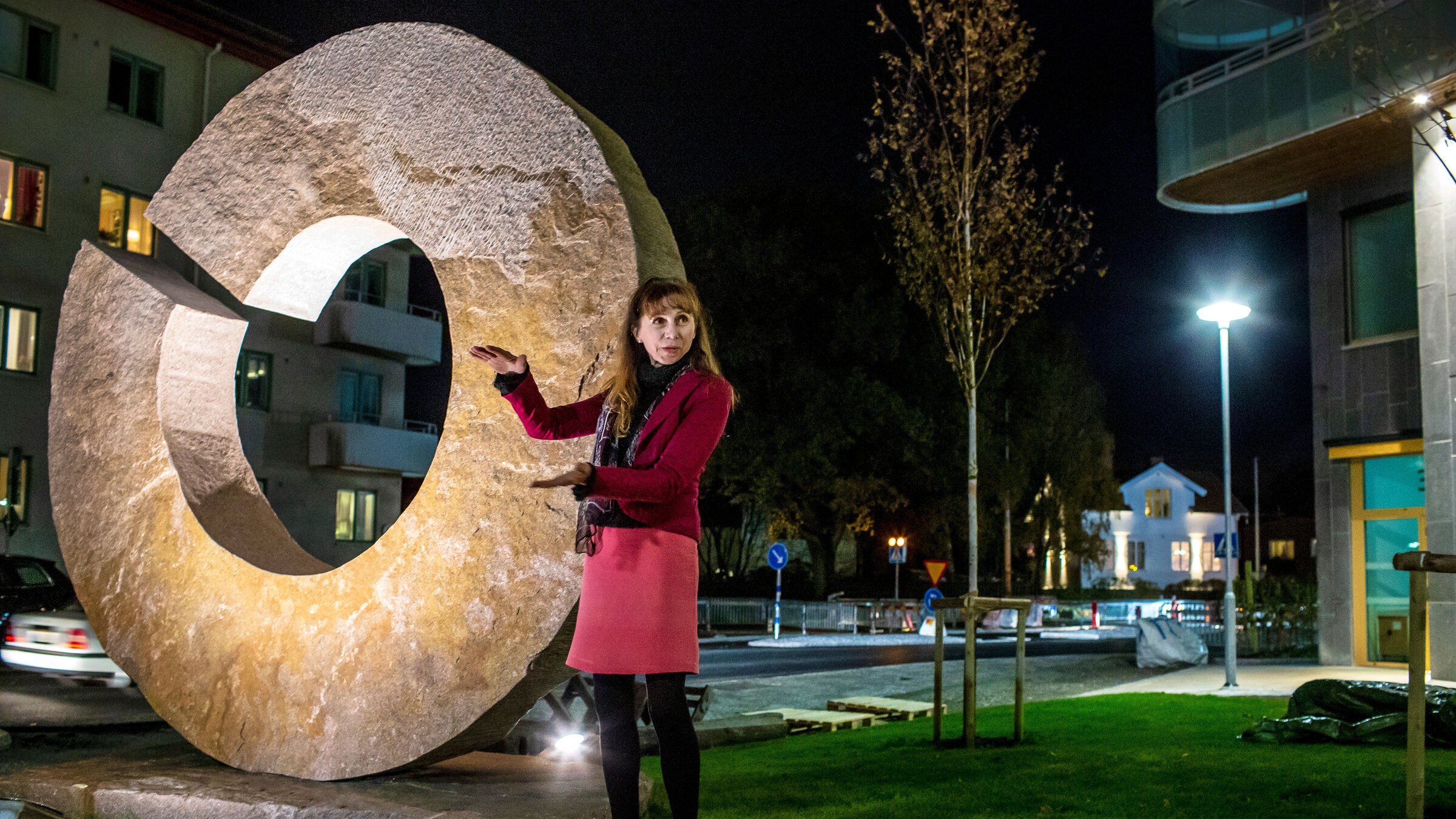 En person i rosa kappa poserar vid en stor stenskulptur på natten i en stadsmiljö.